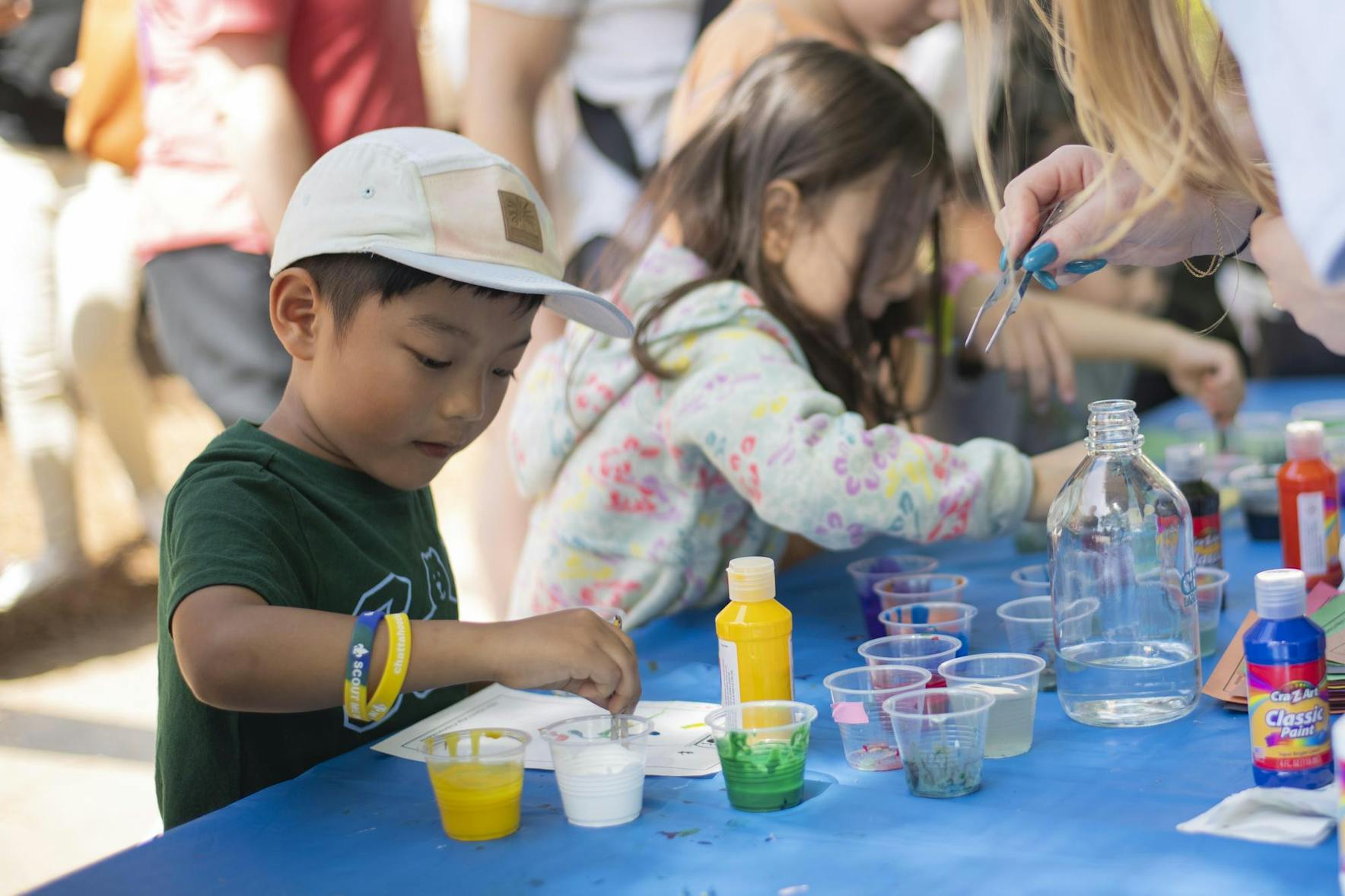 A young child wearing a cap is painting with colorful paints at a table, surrounded by other children and adults.