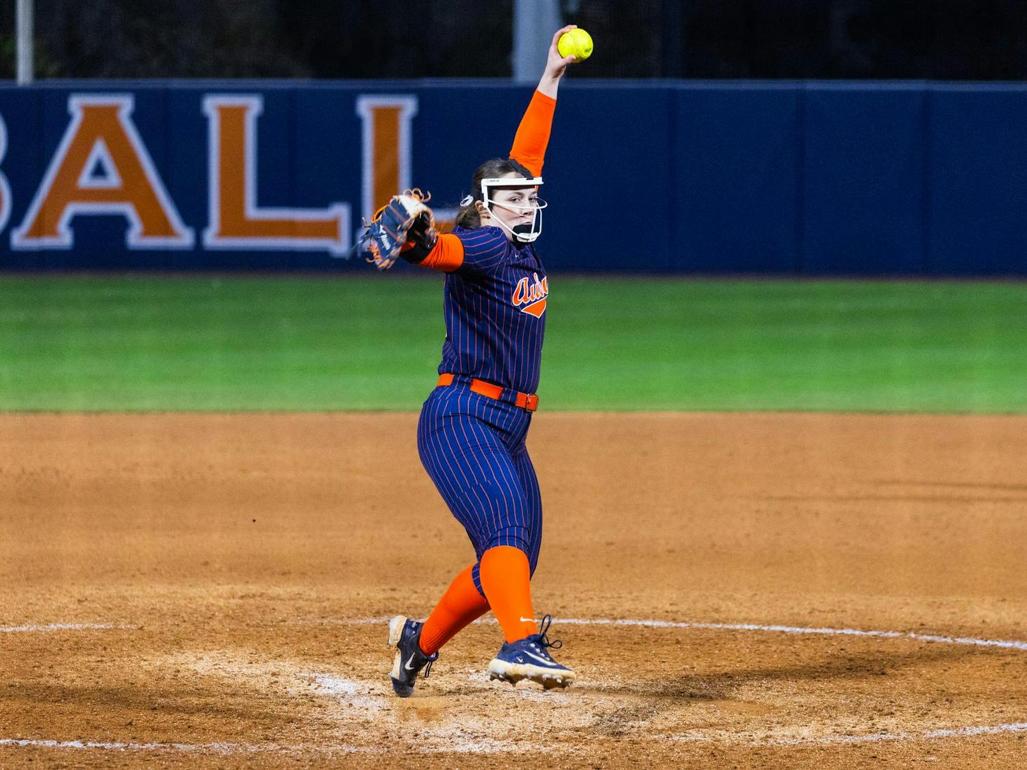 A softball pitcher in a striped uniform winds up with a yellow ball, ready to throw on a dirt field.
