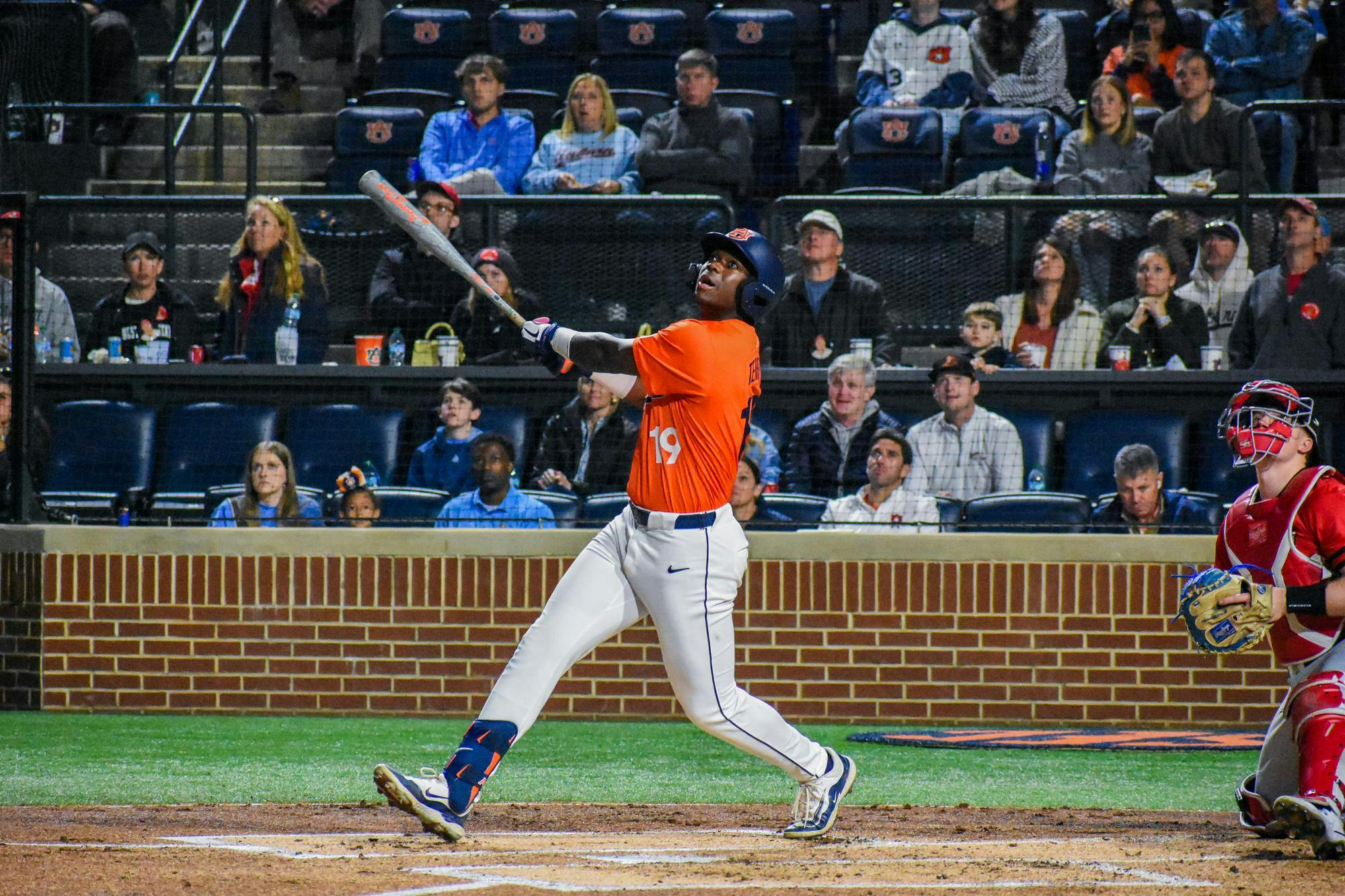 A baseball player in an orange jersey swings a bat, while spectators watch from the stands in the background.