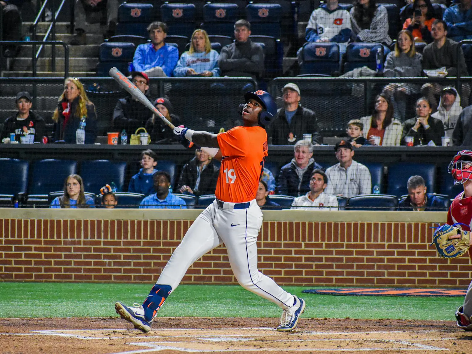 A baseball player in an orange jersey swings a bat, while spectators watch from the stands in the background.