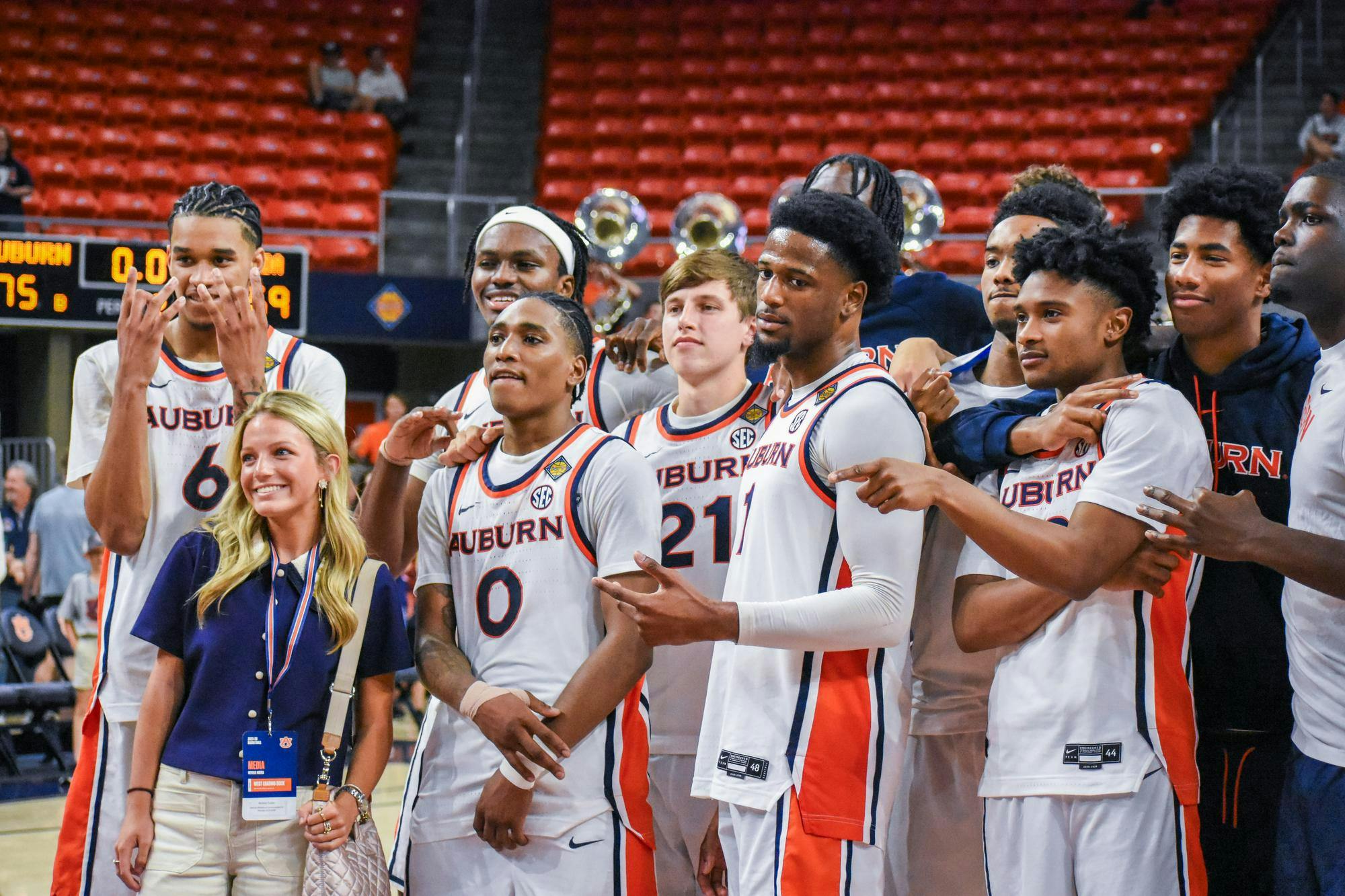 A group of basketball players in uniforms stands together, smiling with a woman positioned in front, all in a sports arena.