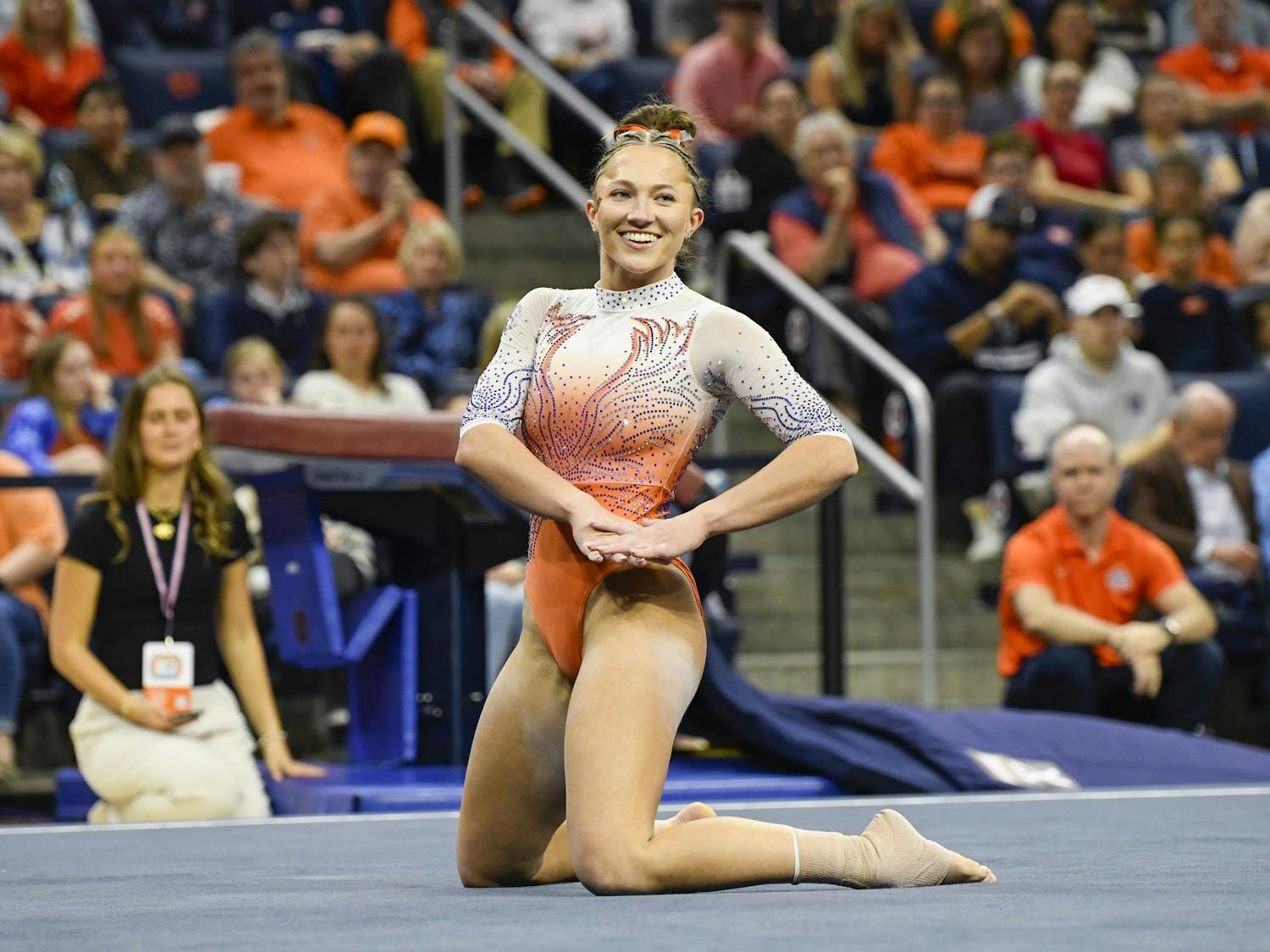 A smiling gymnast in a brightly colored leotard kneels on a mat, surrounded by an audience.