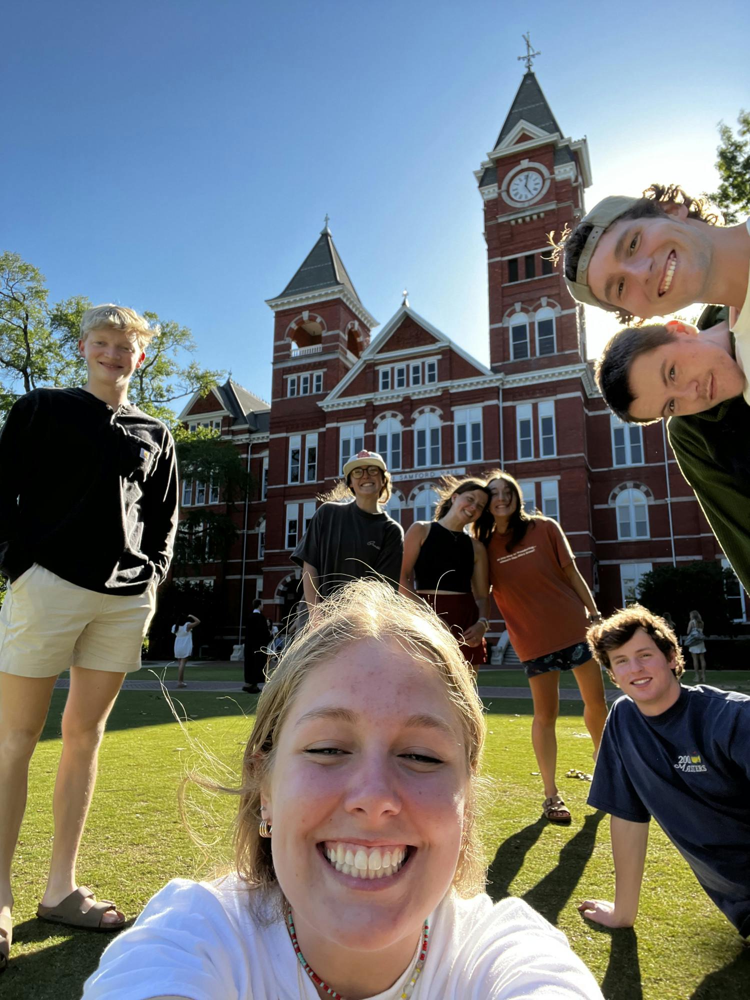 A group of six young adults poses cheerfully on grass in front of a large brick building with a clock tower.