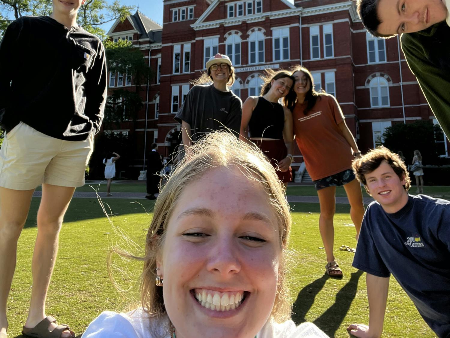 A group of six young adults poses cheerfully on grass in front of a large brick building with a clock tower.