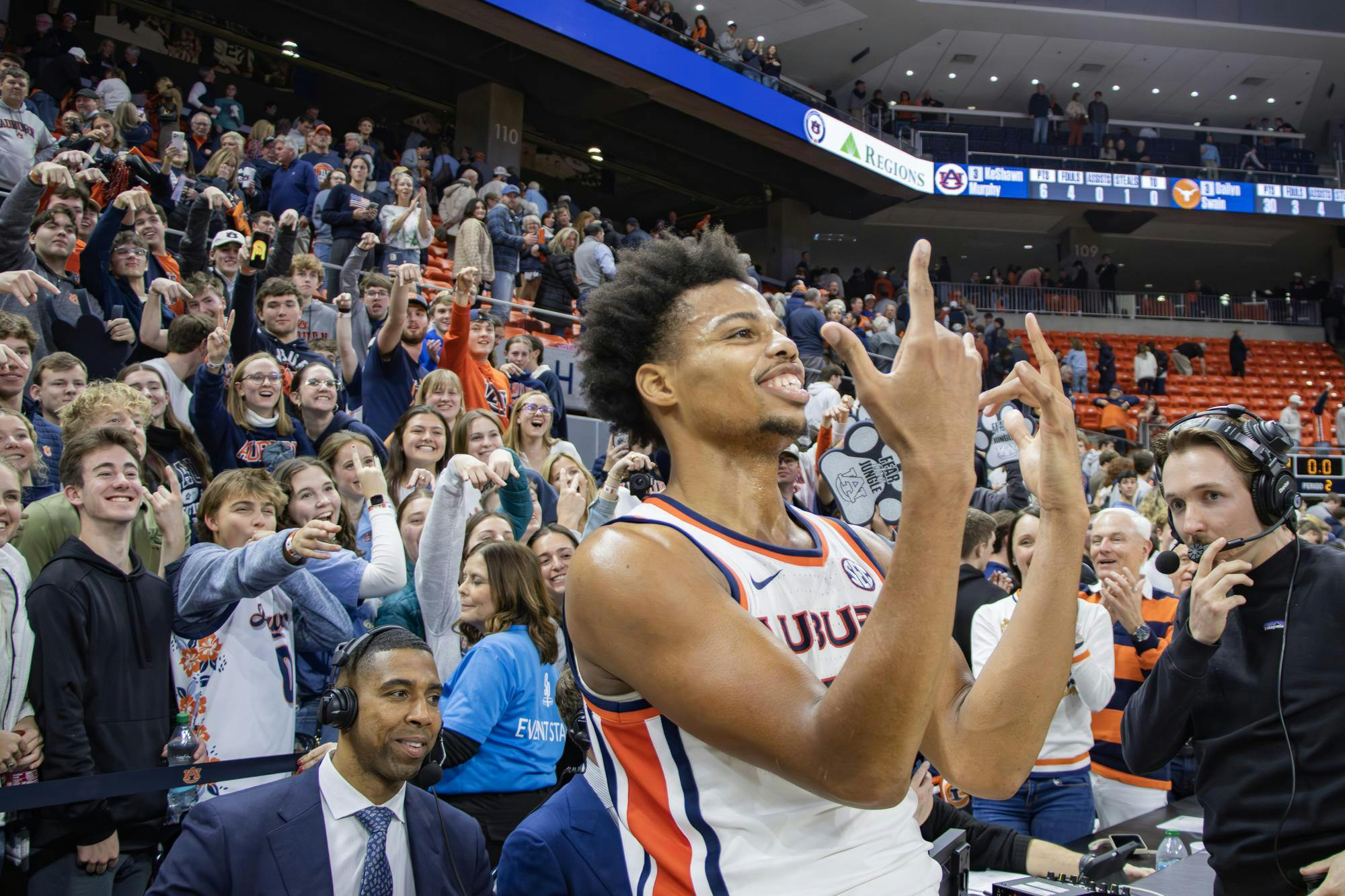 A player in a basketball uniform celebrates enthusiastically while surrounded by a cheering crowd, with commentators nearby.