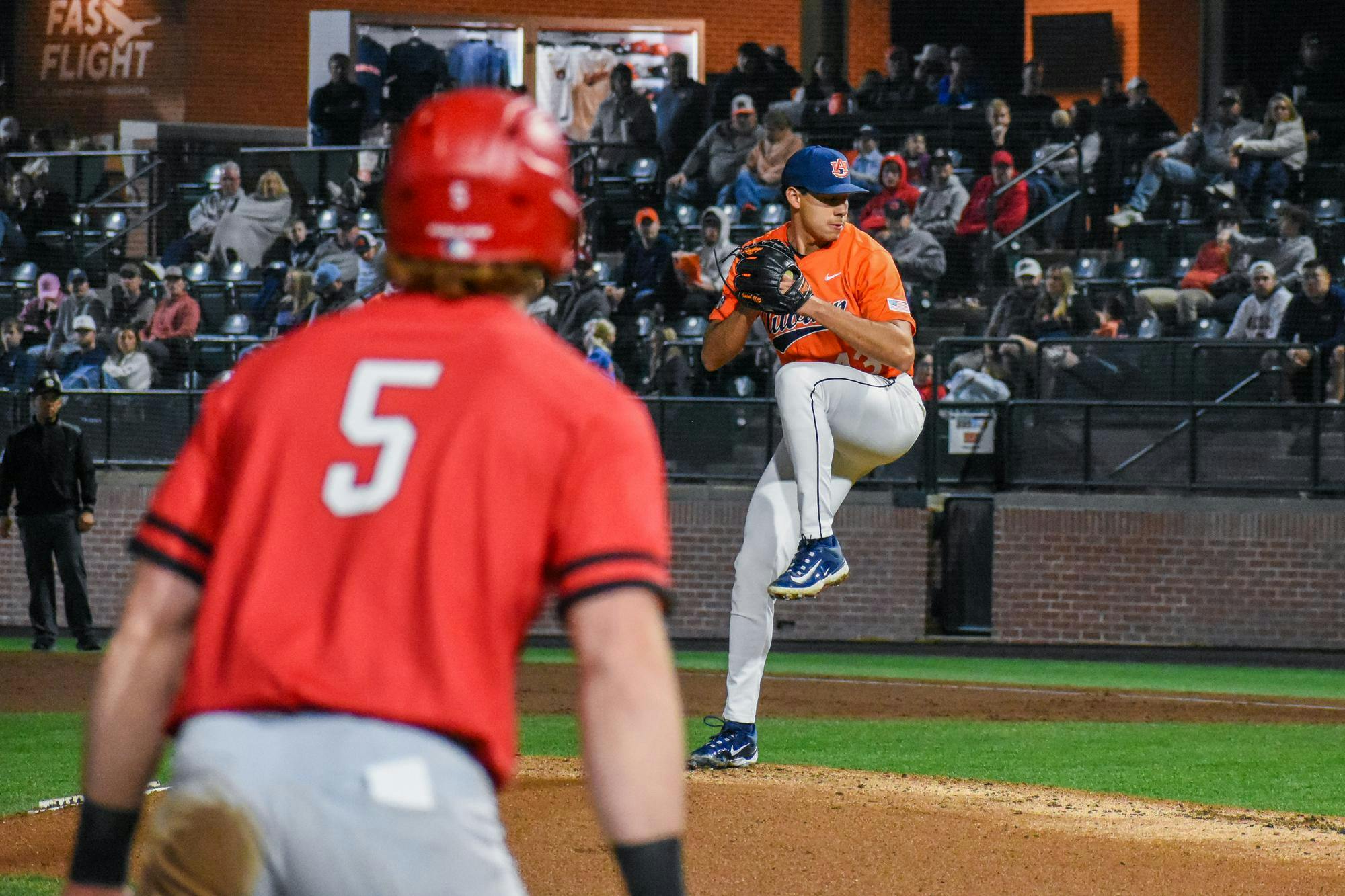 A baseball pitcher in an orange jersey prepares to throw as a batter in a red jersey watches from the plate, with spectators in the background.