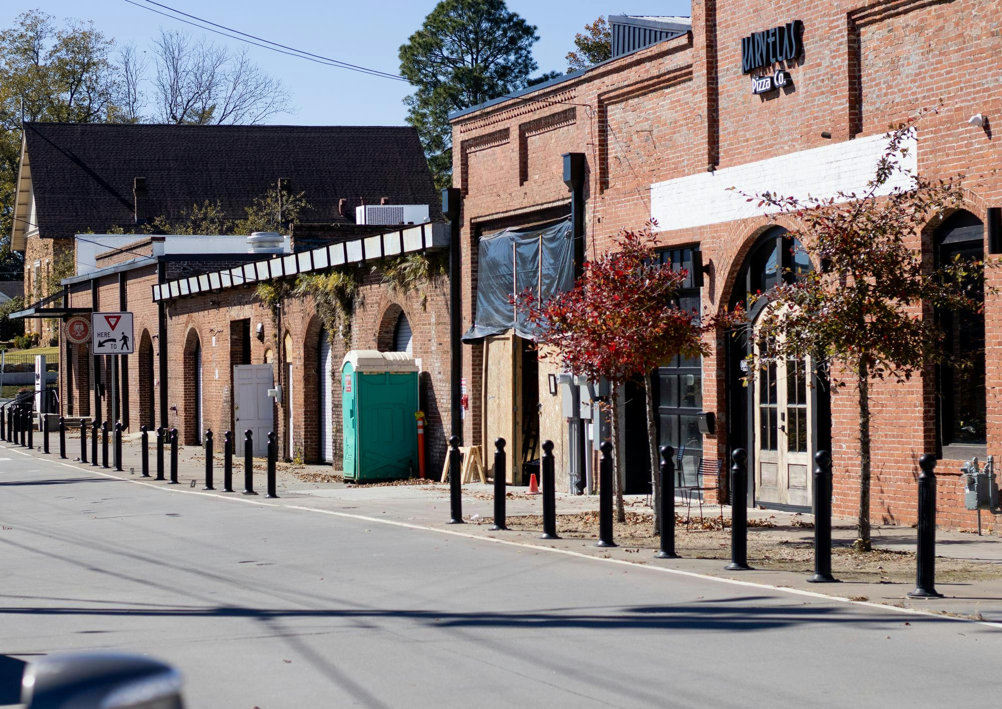 A row of brick buildings with arched windows and a blue portable restroom along a street lined with black bollards.