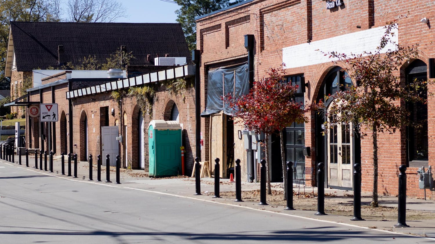 A row of brick buildings with arched windows and a blue portable restroom along a street lined with black bollards.