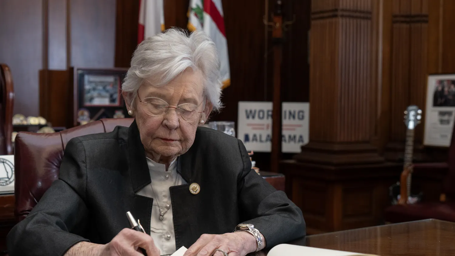 A woman with glasses and gray hair is writing on a document at a polished wooden desk, surrounded by flags and framed photos.