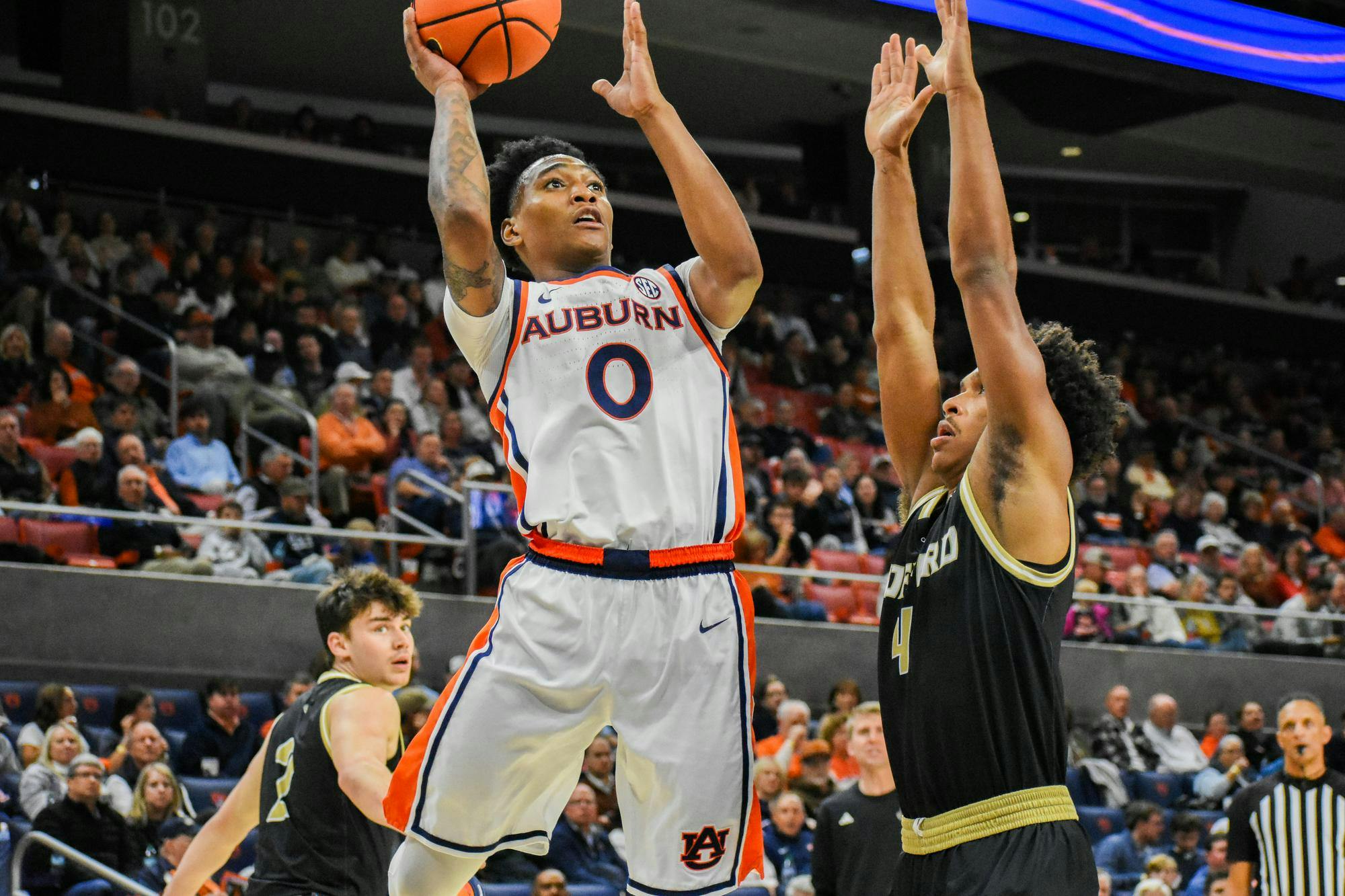 A player in a white and orange uniform is jumping to shoot a basketball while being defended by an opponent in a black uniform.