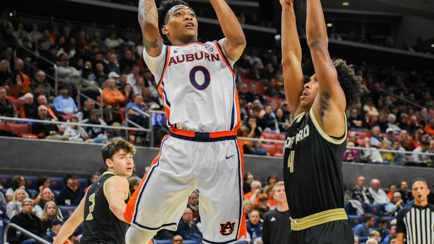 A player in a white and orange uniform is jumping to shoot a basketball while being defended by an opponent in a black uniform.
