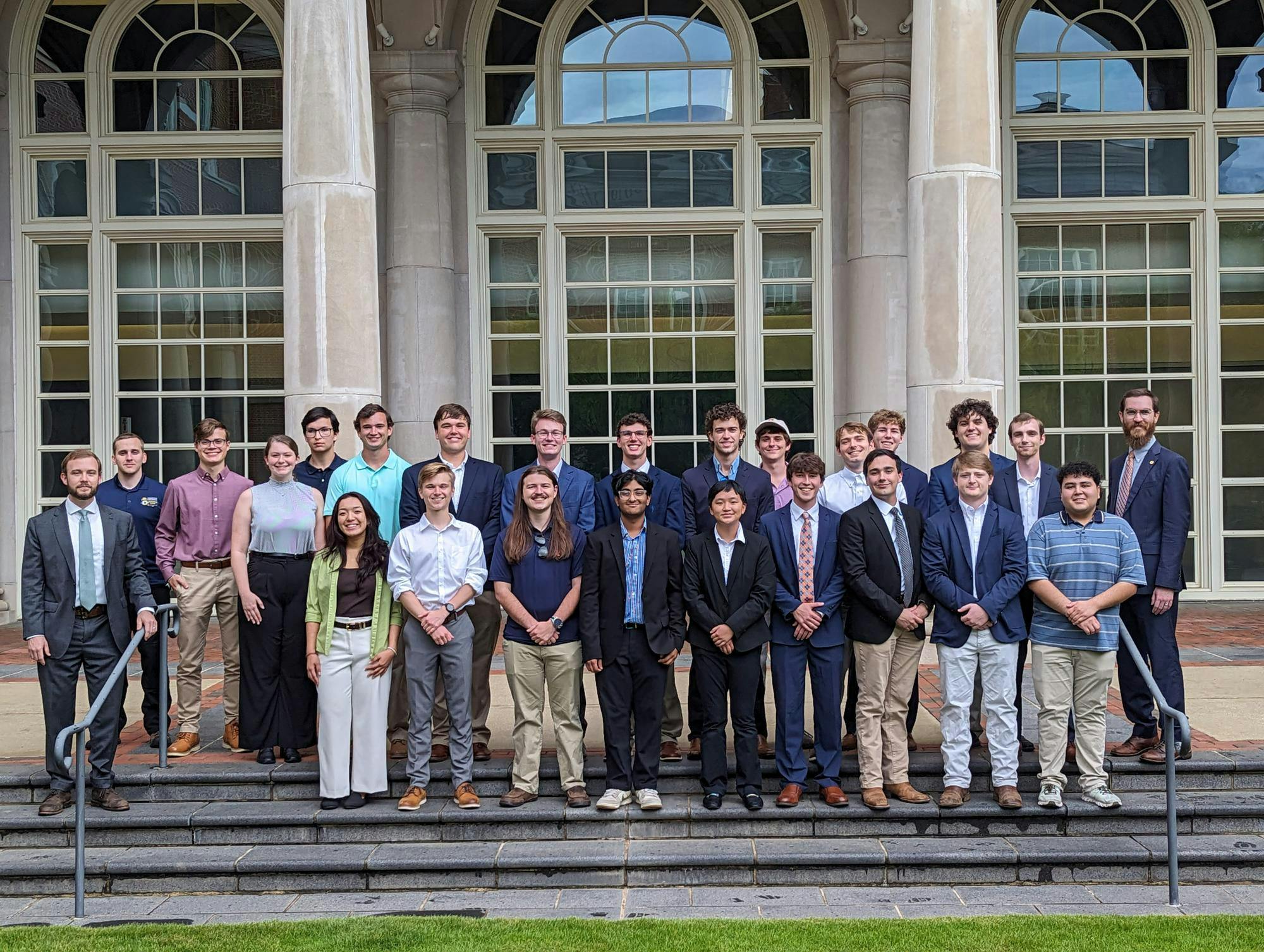 A group of electrical engineering students who participated in the Summer Undergraduate Research Experience, stands on steps in front of large, arched windows, dressed in formal and semi-formal attire.