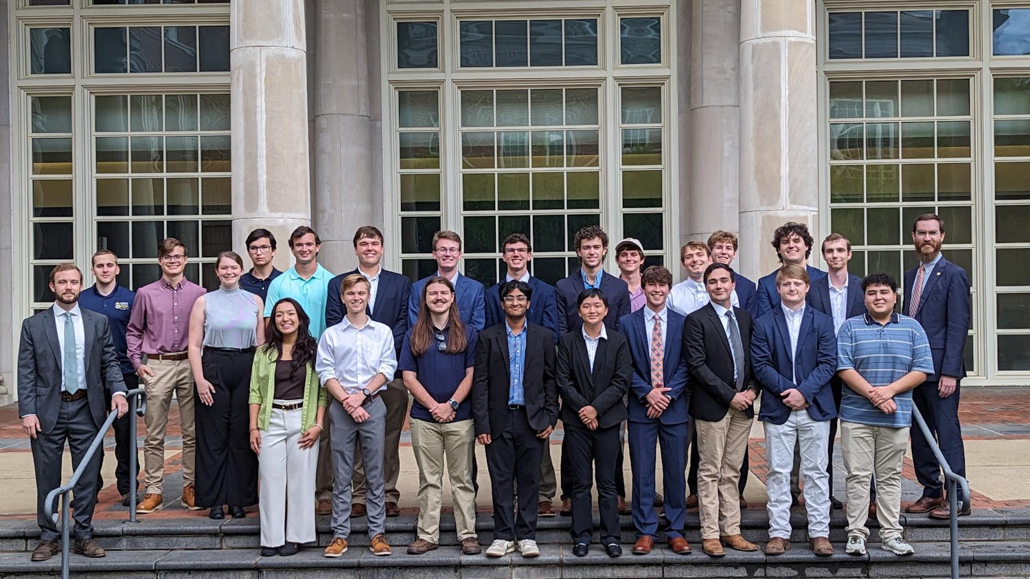A group of electrical engineering students who participated in the Summer Undergraduate Research Experience, stands on steps in front of large, arched windows, dressed in formal and semi-formal attire.