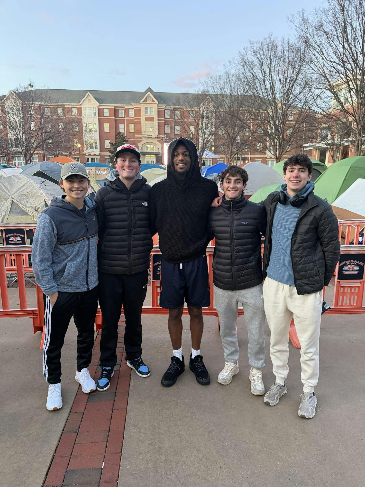 Five young men stand together in front of a row of tents and a nearby building, wearing winter clothing and casual shoes.
