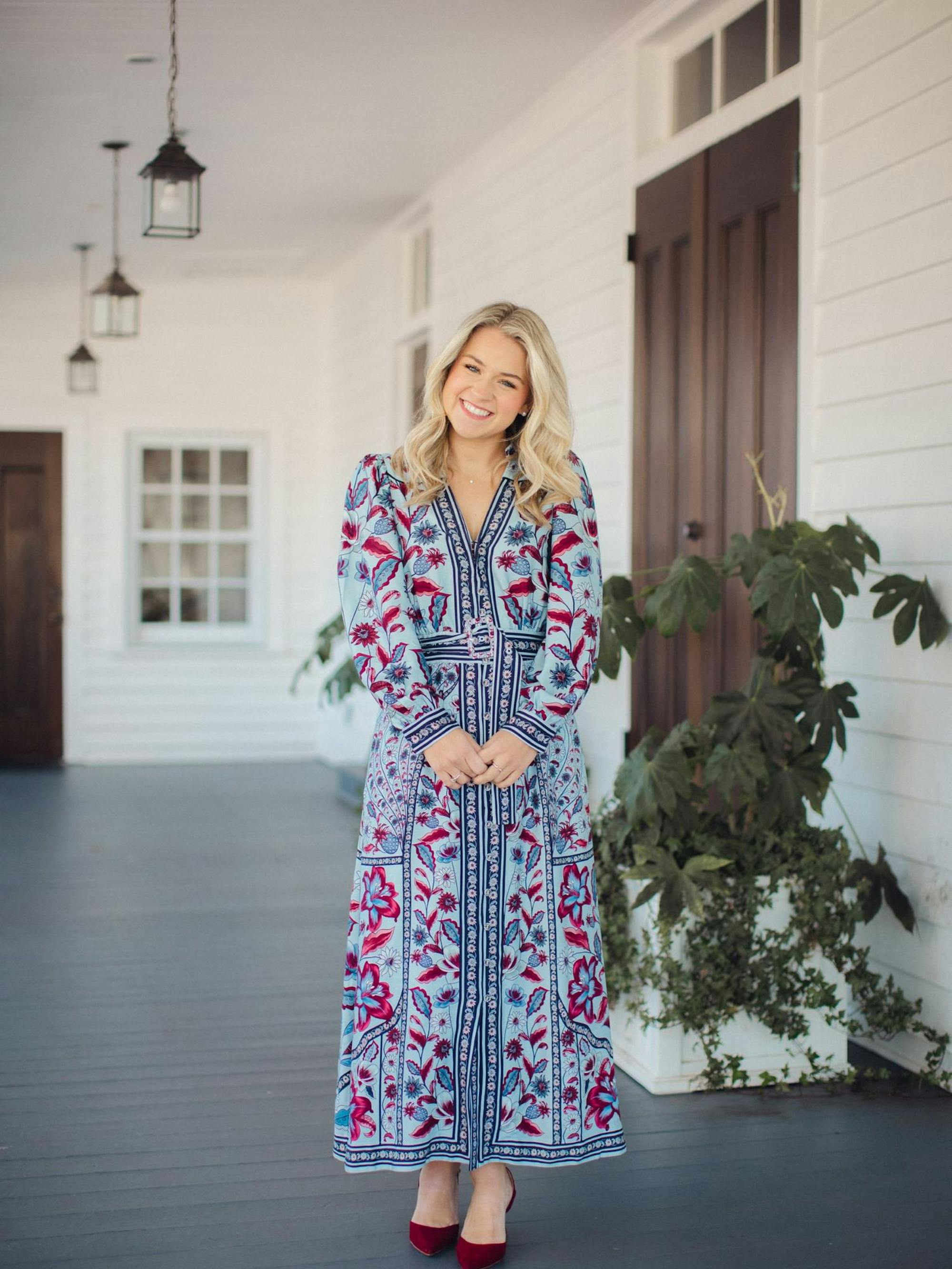 A woman in a floral blue maxi dress stands on a porch, smiling, with potted plants and hanging lanterns around her.