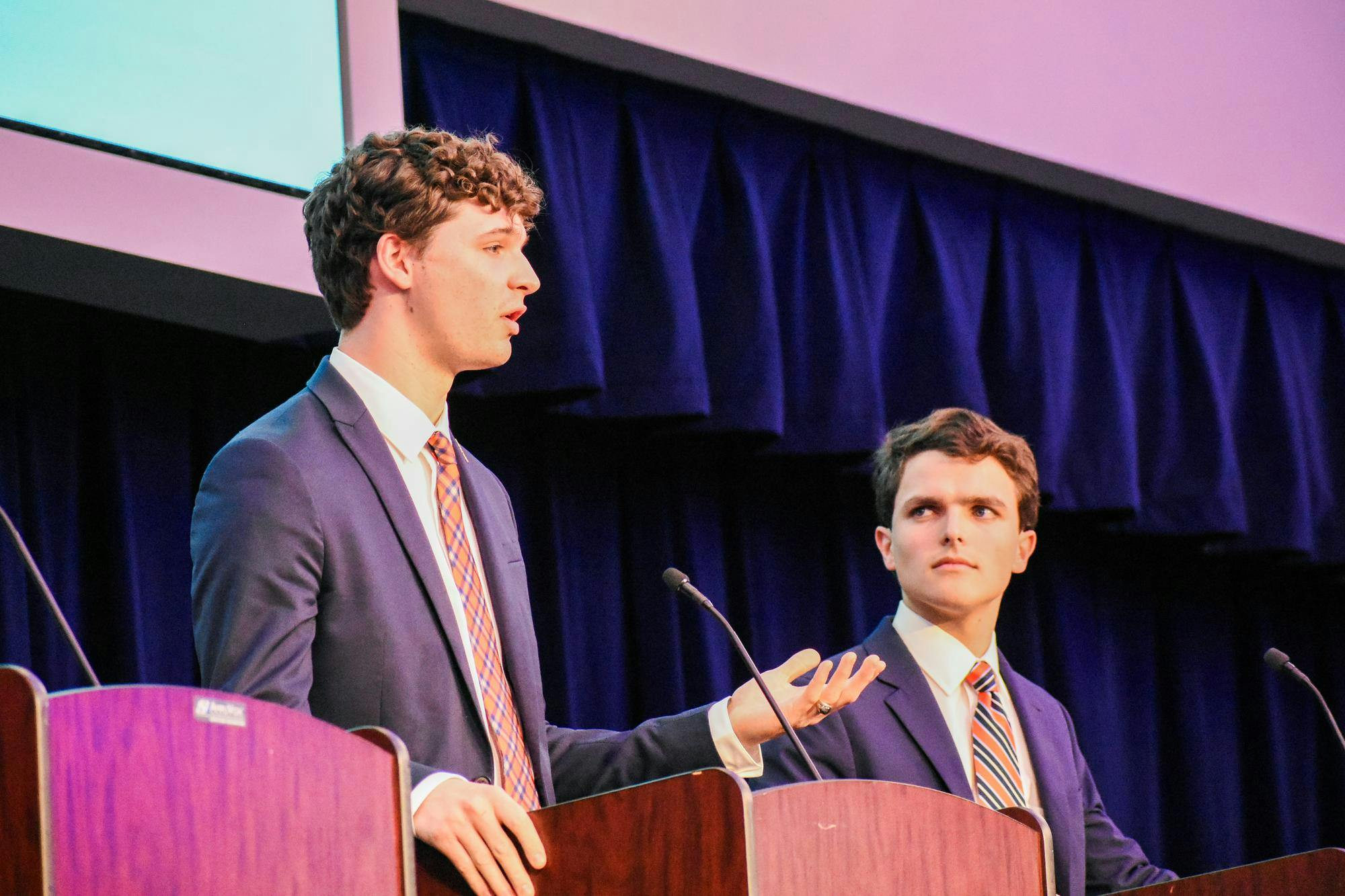 Two young men in suits are engaged in a debate at podiums, with a backdrop of dark fabric and a screen above.