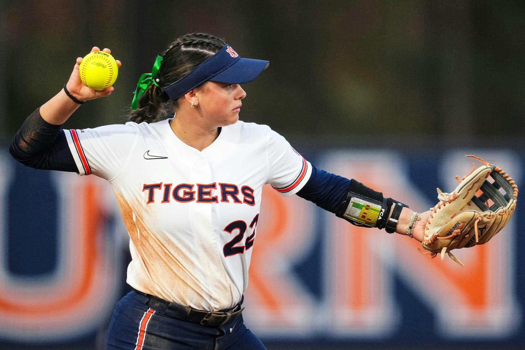 A young woman in a white and blue softball uniform prepares to throw a yellow softball while wearing a glove.