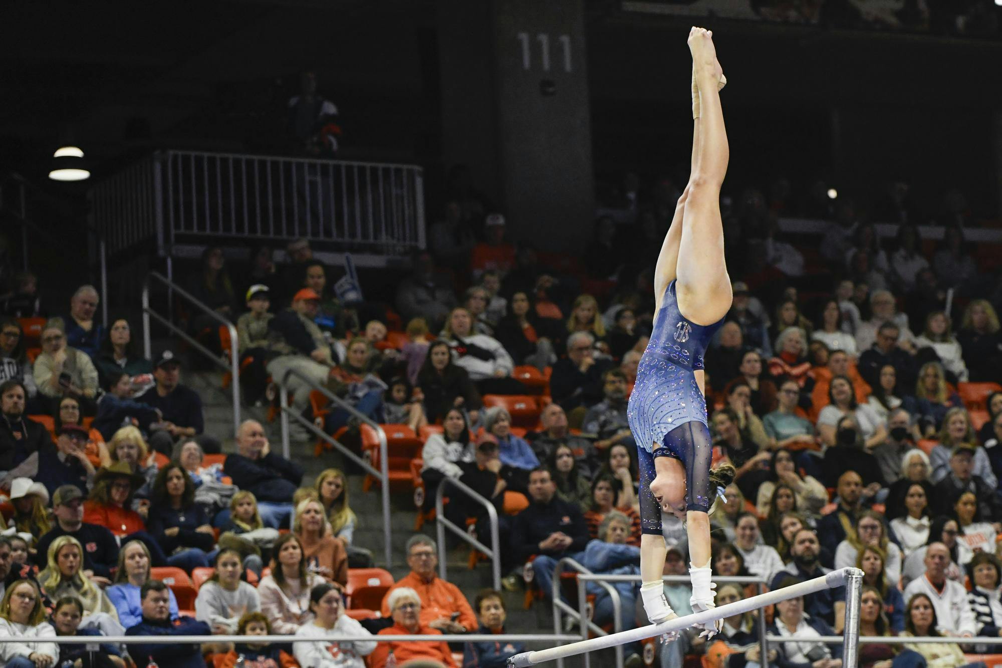 A gymnast performs a handstand on a balance beam in front of a large audience.