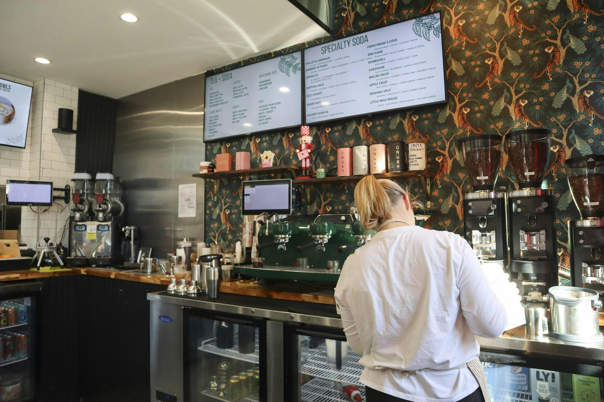 A barista wearing a white shirt prepares drinks at a coffee counter adorned with menus and colorful decor.