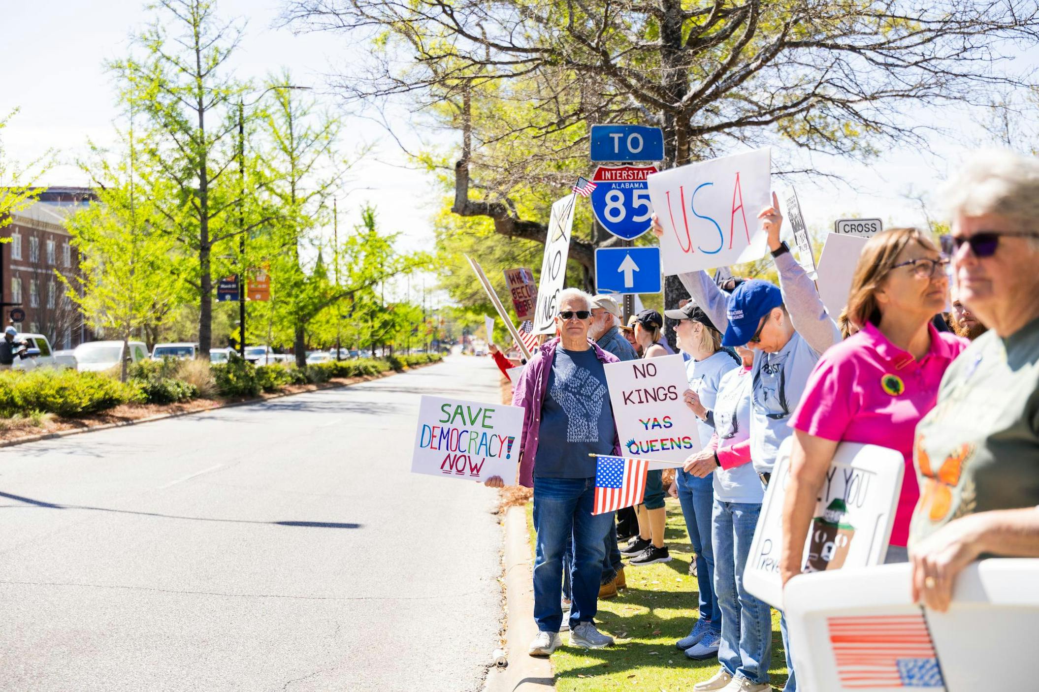 A group of people stands along a street holding various protest signs and flags, with trees and buildings in the background.