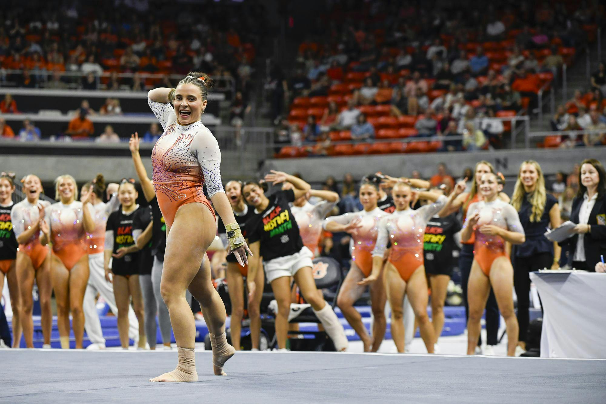 A gymnast in an orange and silver leotard smiles and poses confidently, surrounded by teammates cheering enthusiastically in a sports arena.