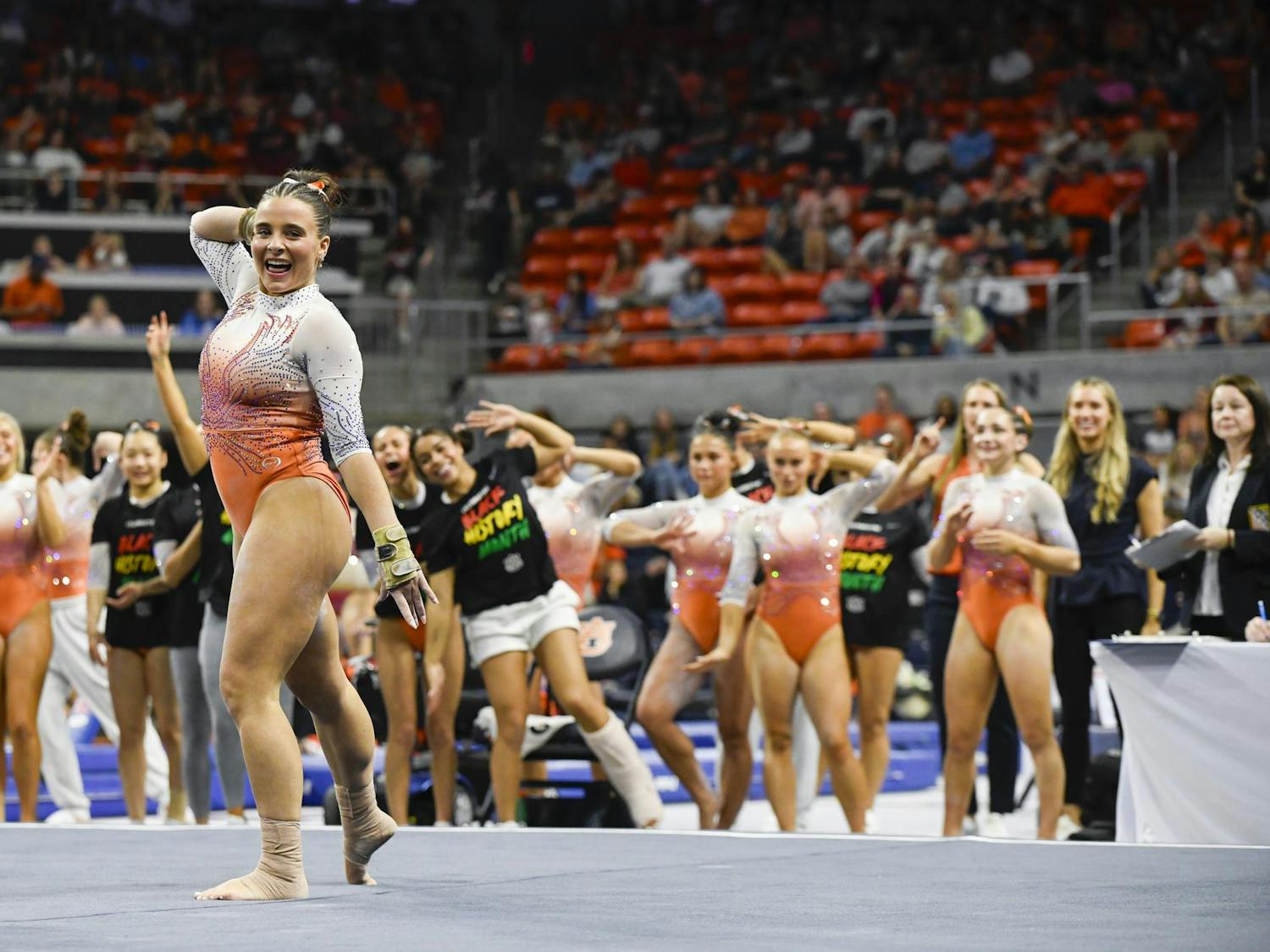 A gymnast in an orange and silver leotard smiles and poses confidently, surrounded by teammates cheering enthusiastically in a sports arena.
