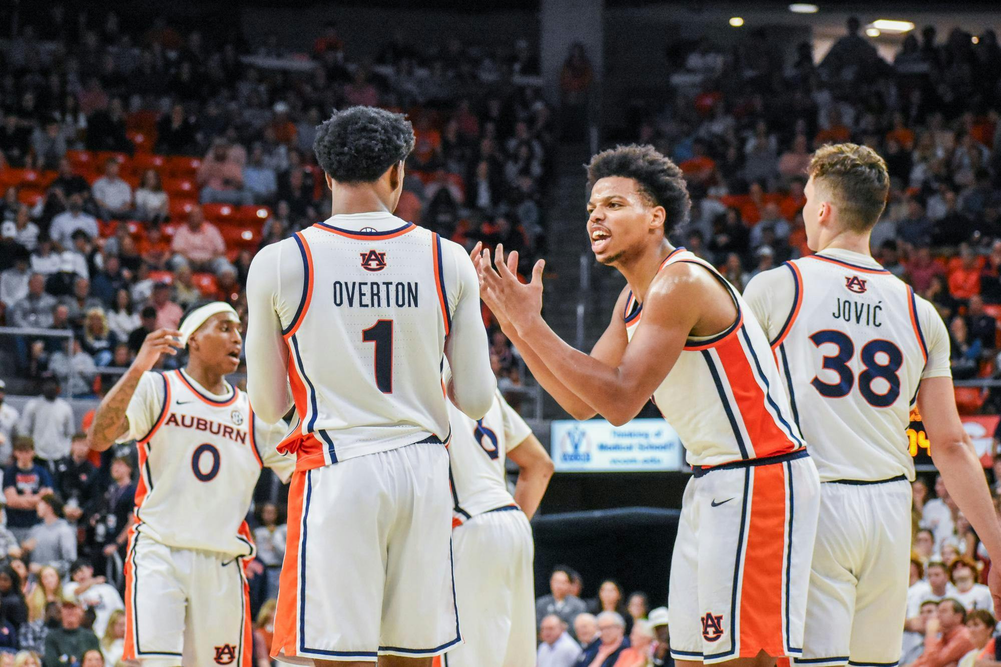 Five basketball players in white uniforms with orange and blue accents are engaged in a discussion on the court.