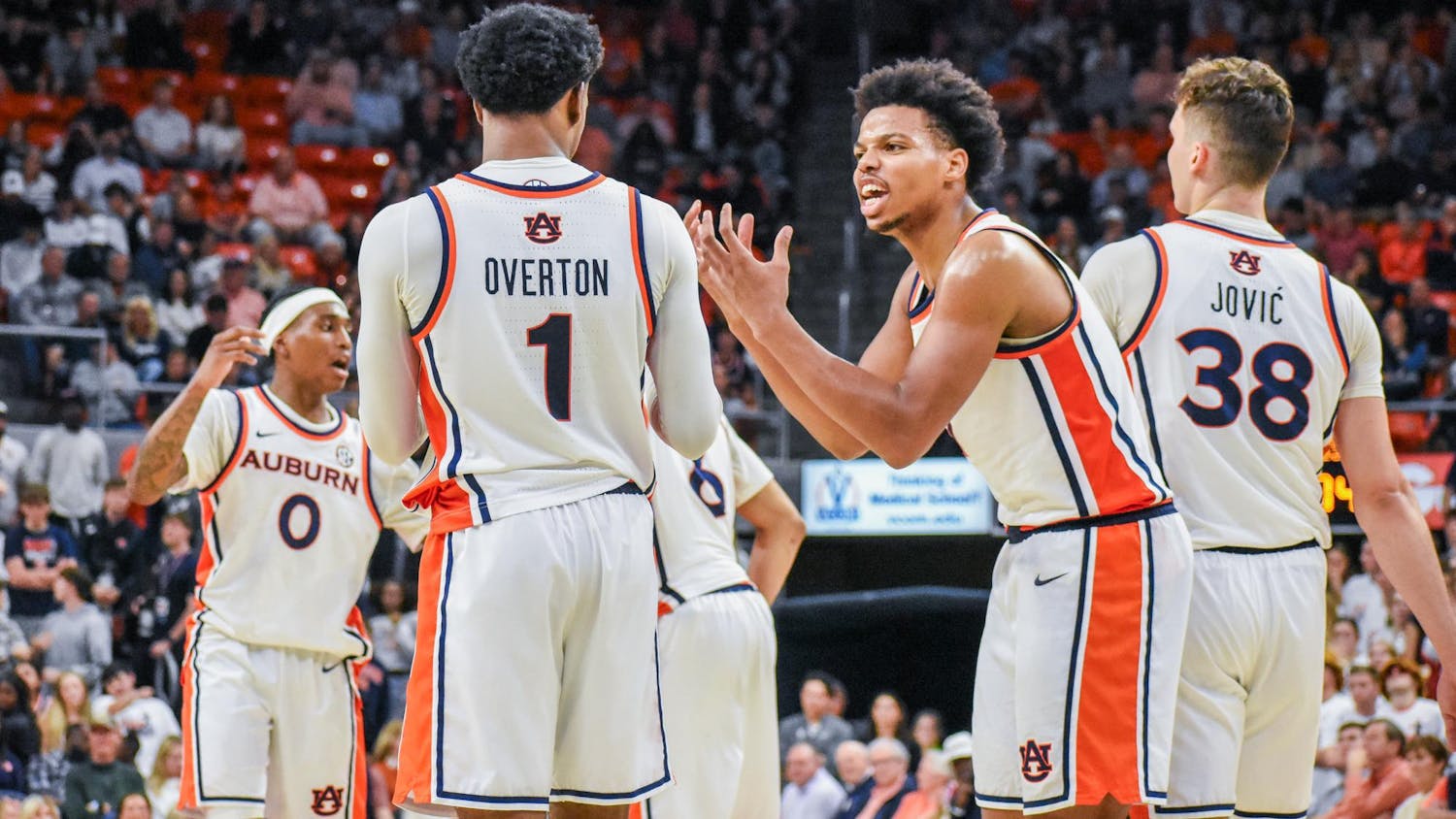 Five basketball players in white uniforms with orange and blue accents are engaged in a discussion on the court.