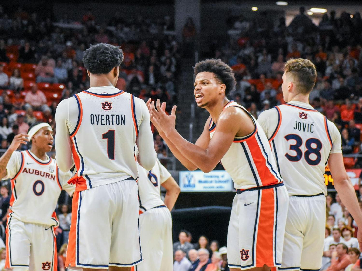 Five basketball players in white uniforms with orange and blue accents are engaged in a discussion on the court.