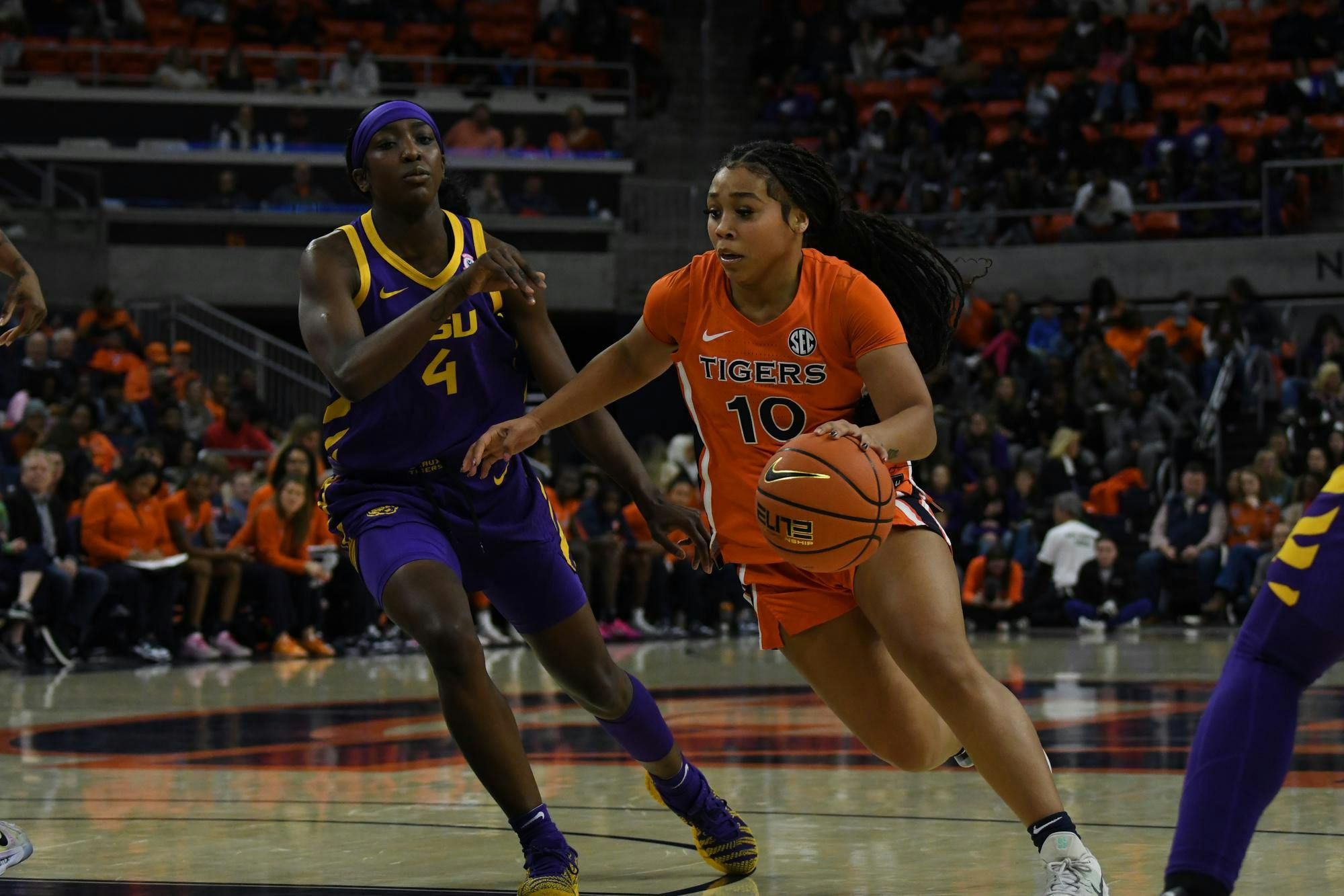 A player in an orange basketball uniform dribbles past an opponent in a purple uniform, with a crowd visible in the background.