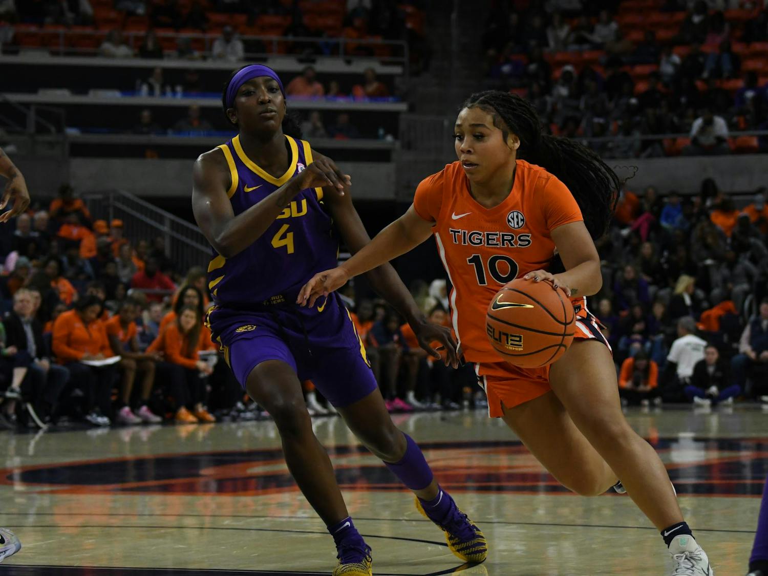 A player in an orange basketball uniform dribbles past an opponent in a purple uniform, with a crowd visible in the background.