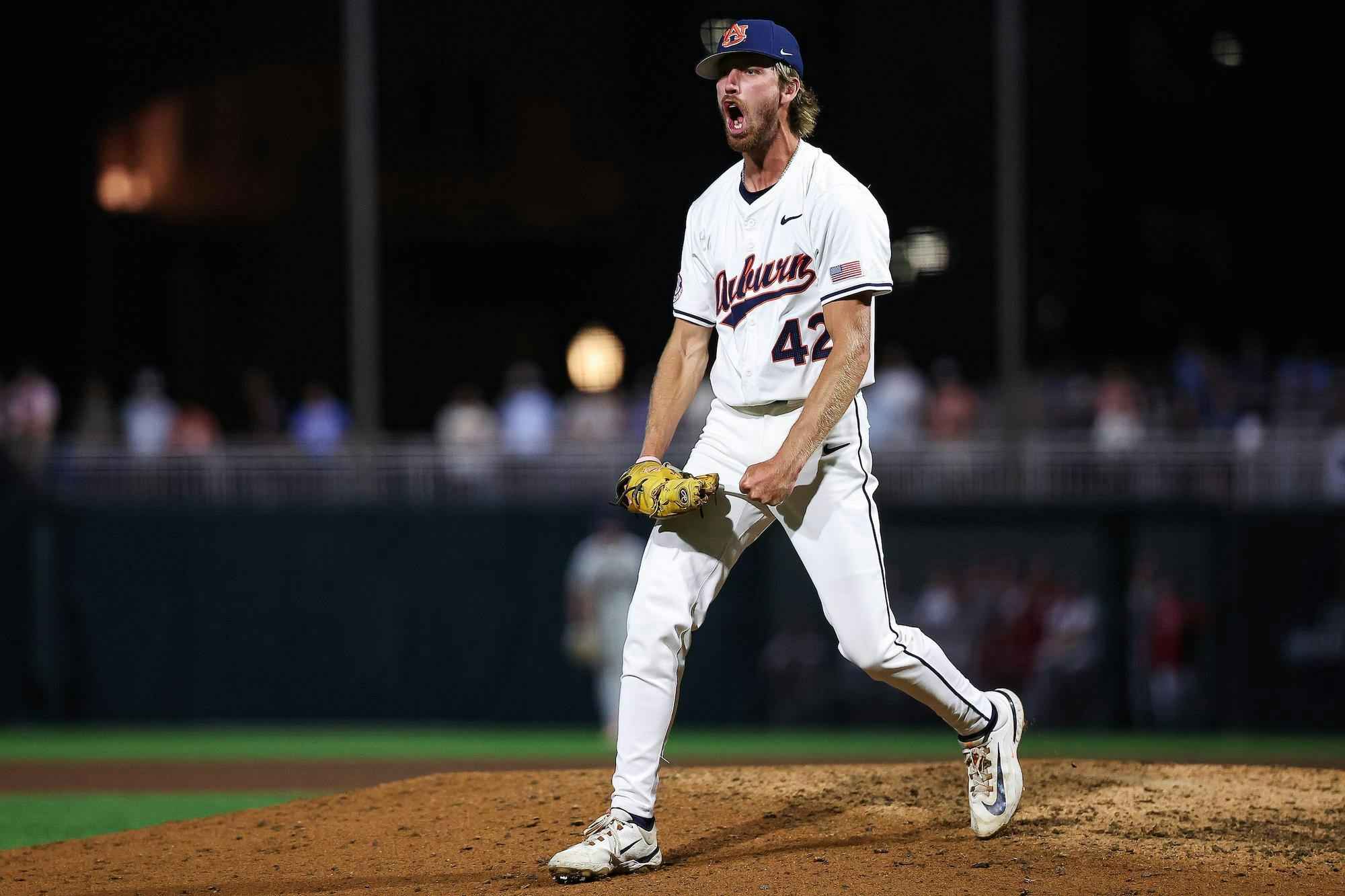 A baseball player wearing a white uniform with blue accents passionately celebrates on the pitcher's mound at night.