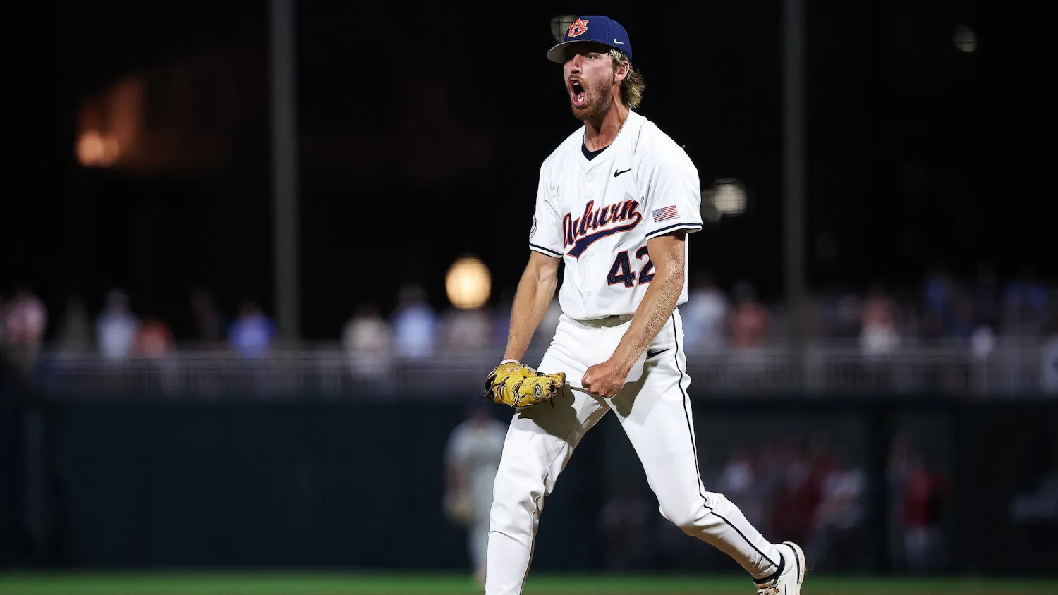 A baseball player wearing a white uniform with blue accents passionately celebrates on the pitcher's mound at night.