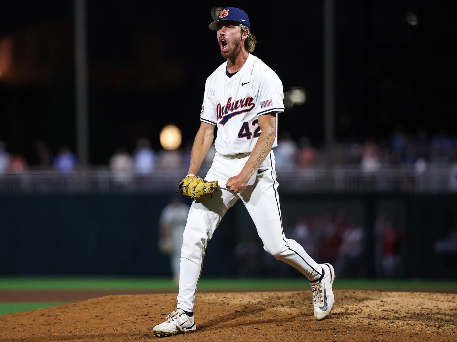 A baseball player wearing a white uniform with blue accents passionately celebrates on the pitcher's mound at night.