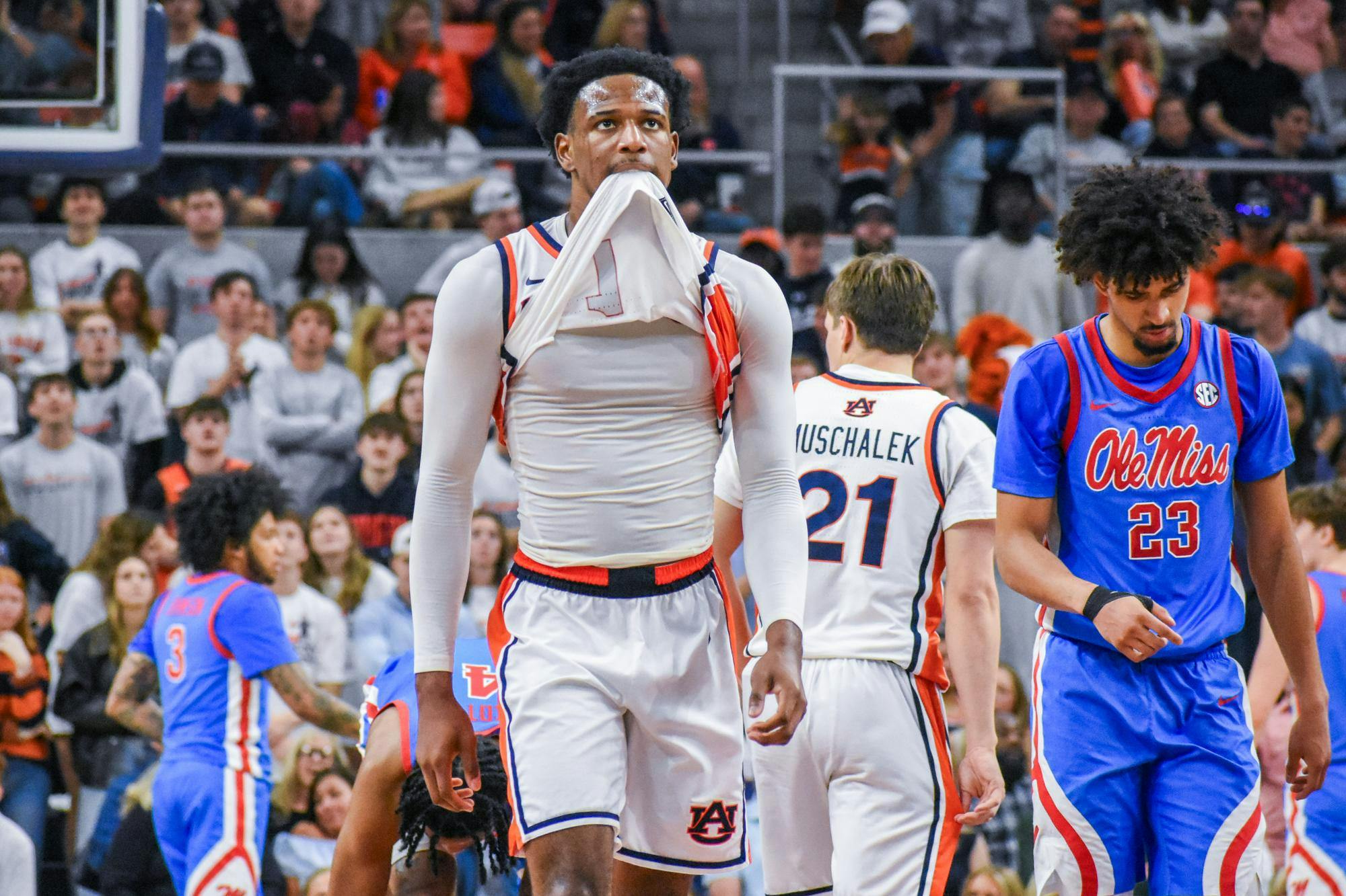 A basketball player in a white jersey pulls a shirt over his mouth while other players and a cheering crowd are in the background.