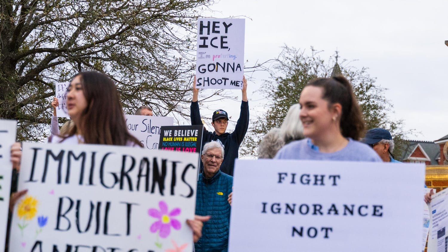A diverse group of protesters hold signs advocating for immigration rights and social justice while standing under a cloudy sky.