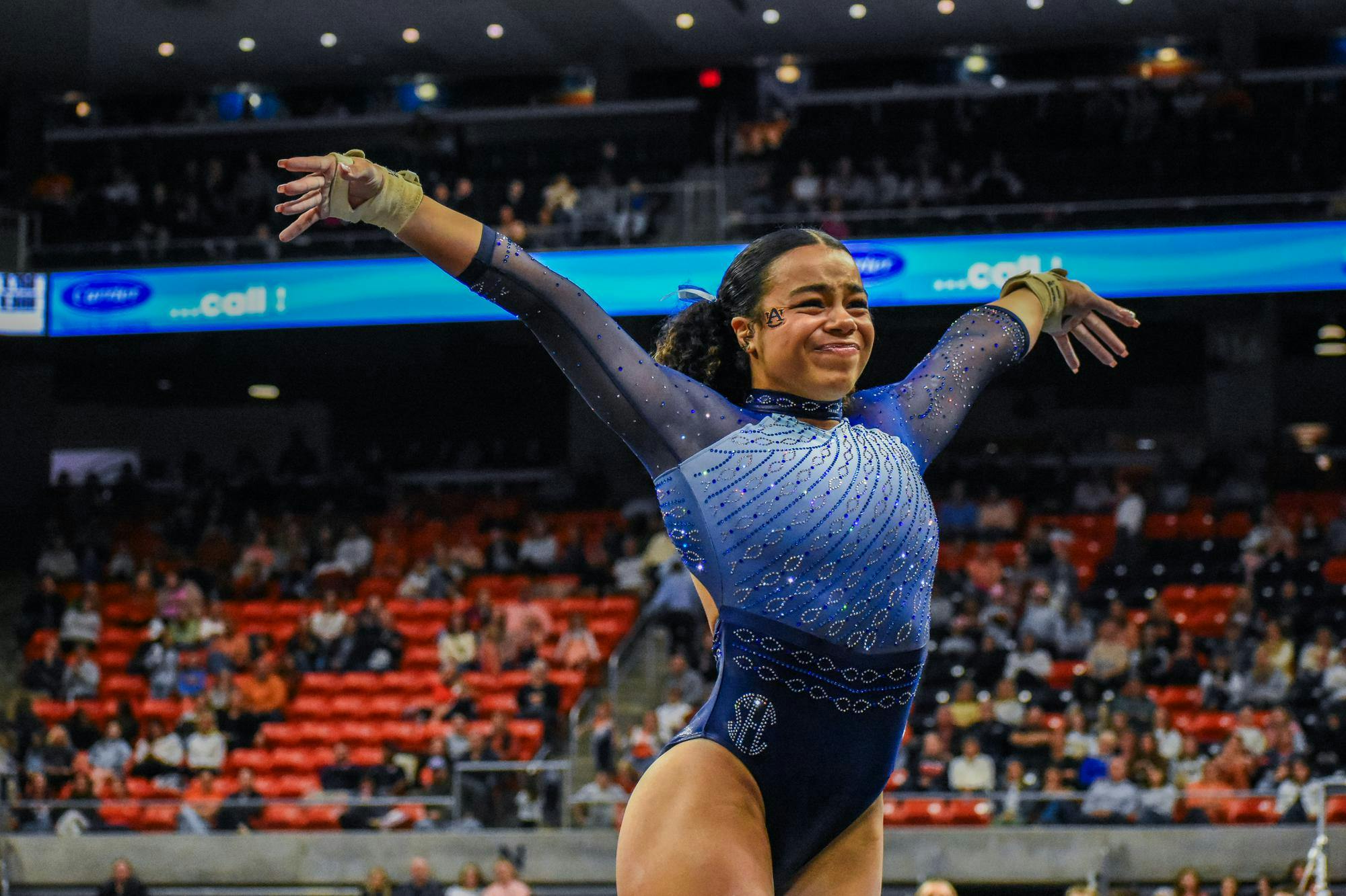 A gymnast with a beaming smile performs in a sparkling blue outfit, arms outstretched, in front of a cheering crowd.