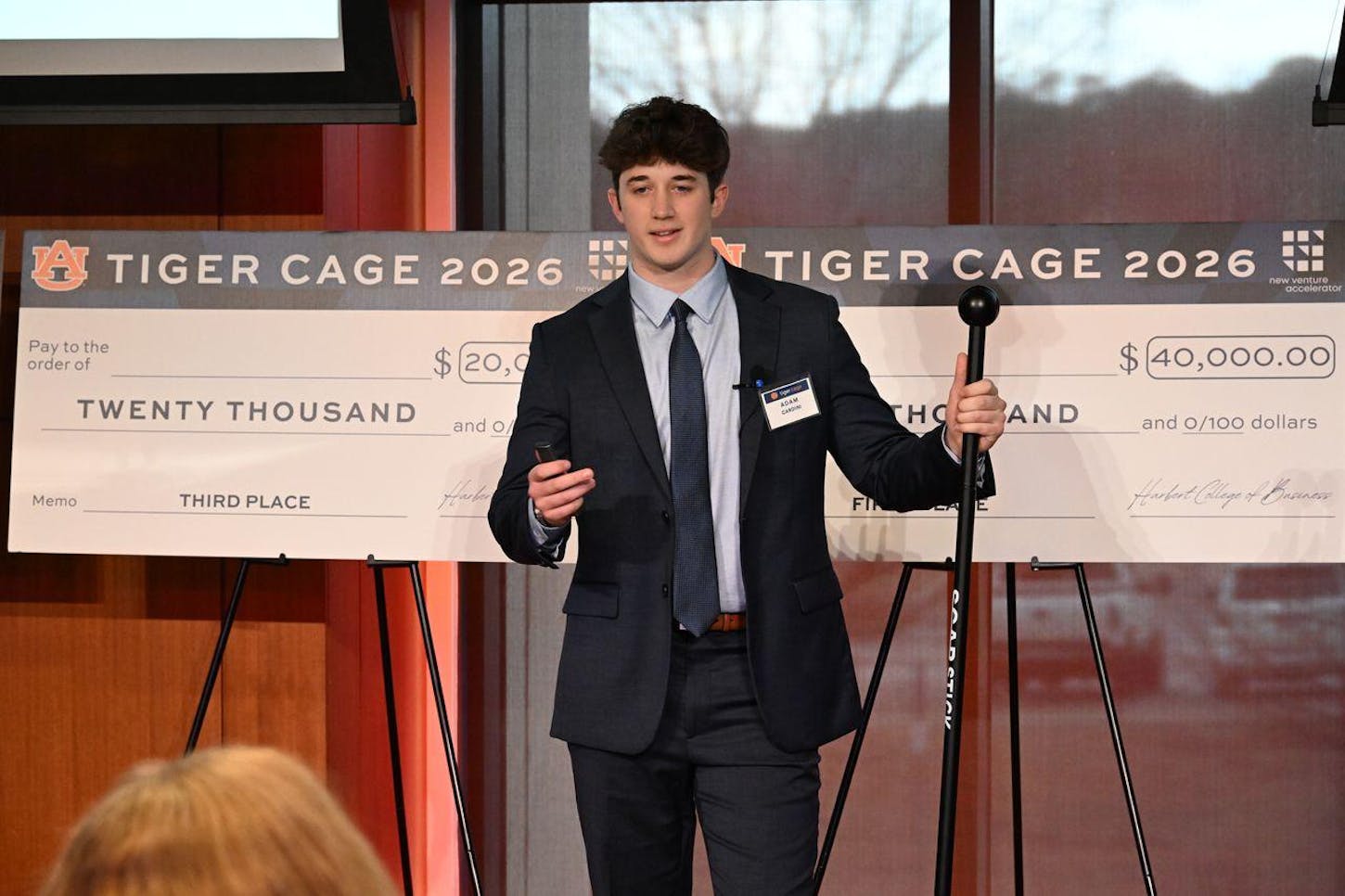 A young man in a suit presents beside a large check labeled for twenty thousand dollars and marked third place.