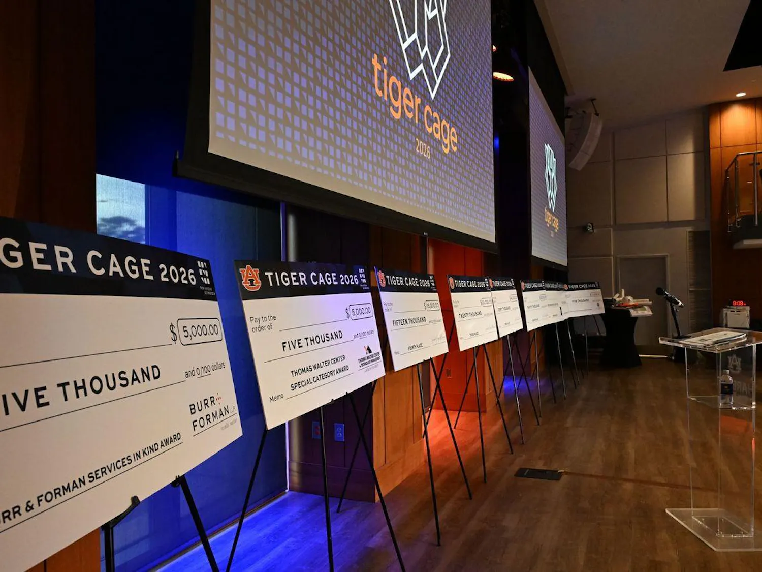 Rows of oversized novelty checks display various amounts and awards against a backdrop featuring a logo for "Tiger Cage 2026."