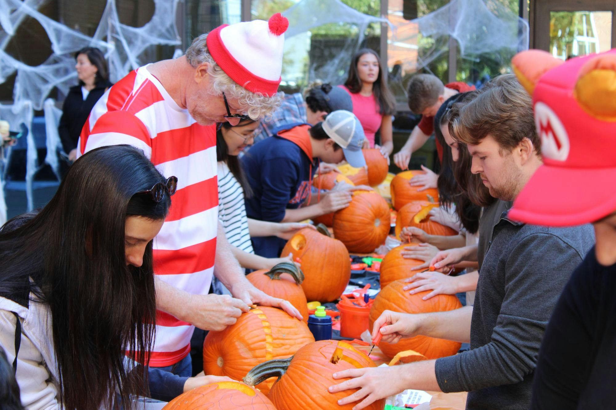 People gather around a table, carving pumpkins and decorating them, with a backdrop of spider webs.