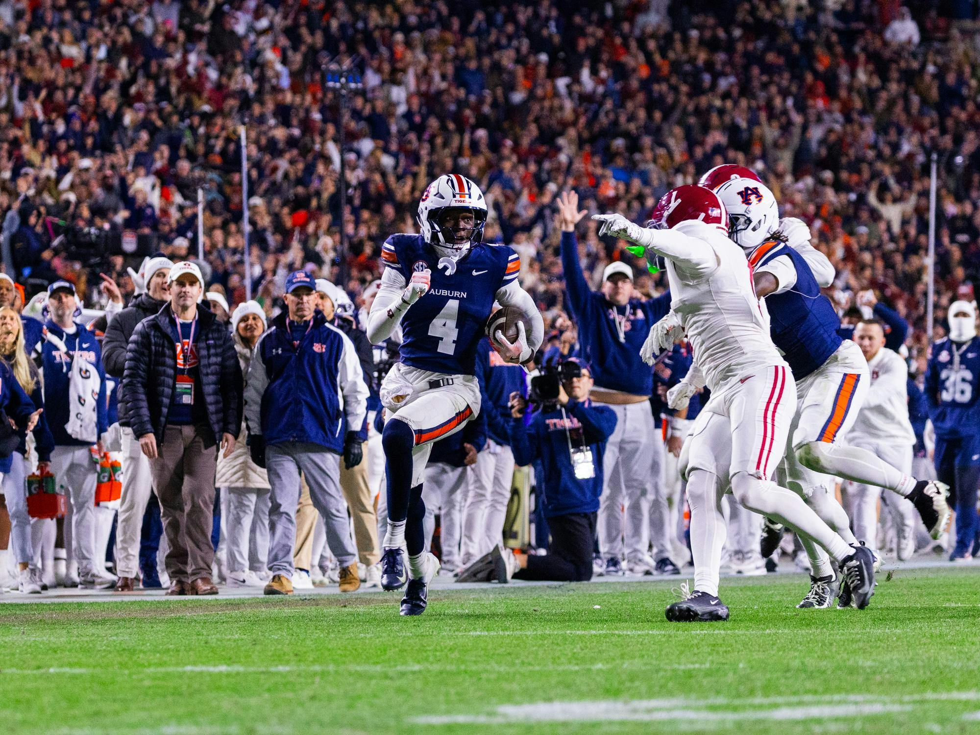 A football player in a blue uniform evades defenders while running towards the sidelines, with a cheering crowd in the background.