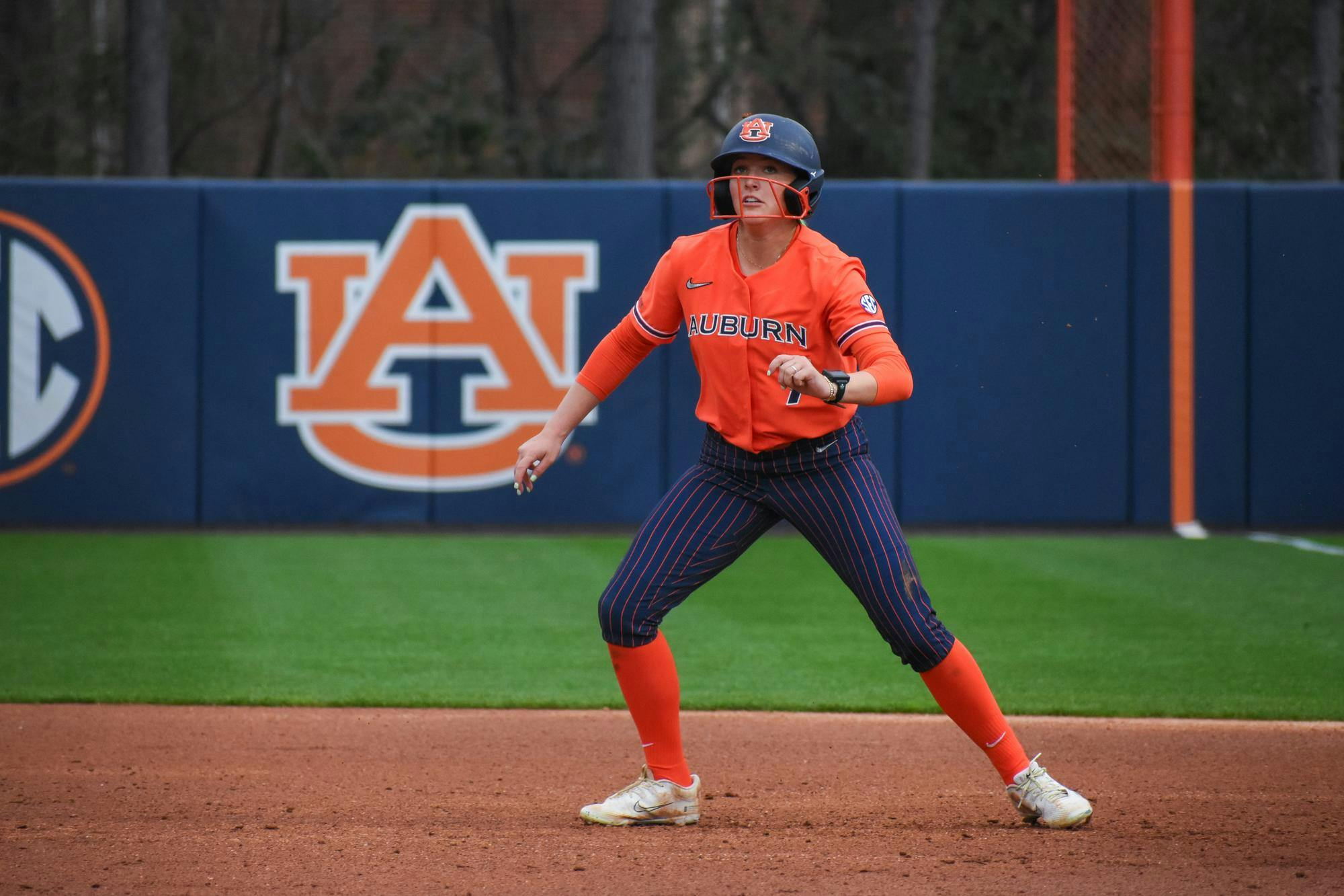 A female softball player in an orange uniform with blue stripes stands on a dirt field, poised and focused on the game.