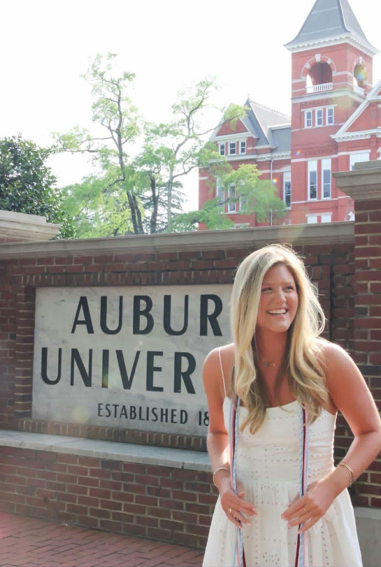 A woman in a white dress stands smiling in front of a brick sign that reads "AUBURN UNIVERSITY."