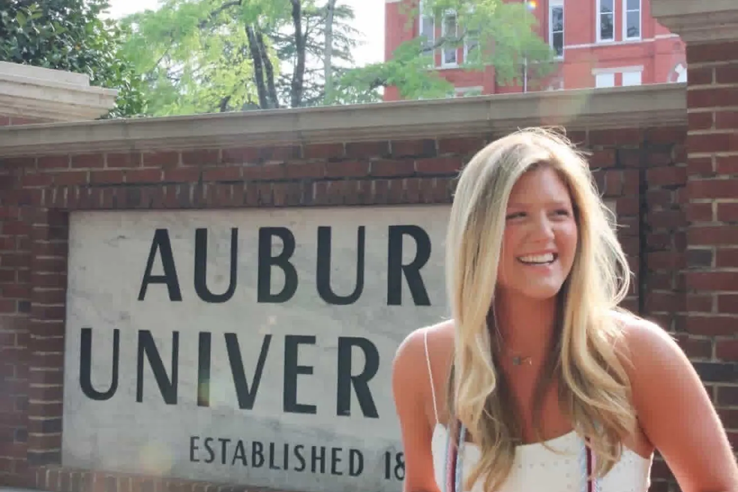 A woman in a white dress stands smiling in front of a brick sign that reads "AUBURN UNIVERSITY."