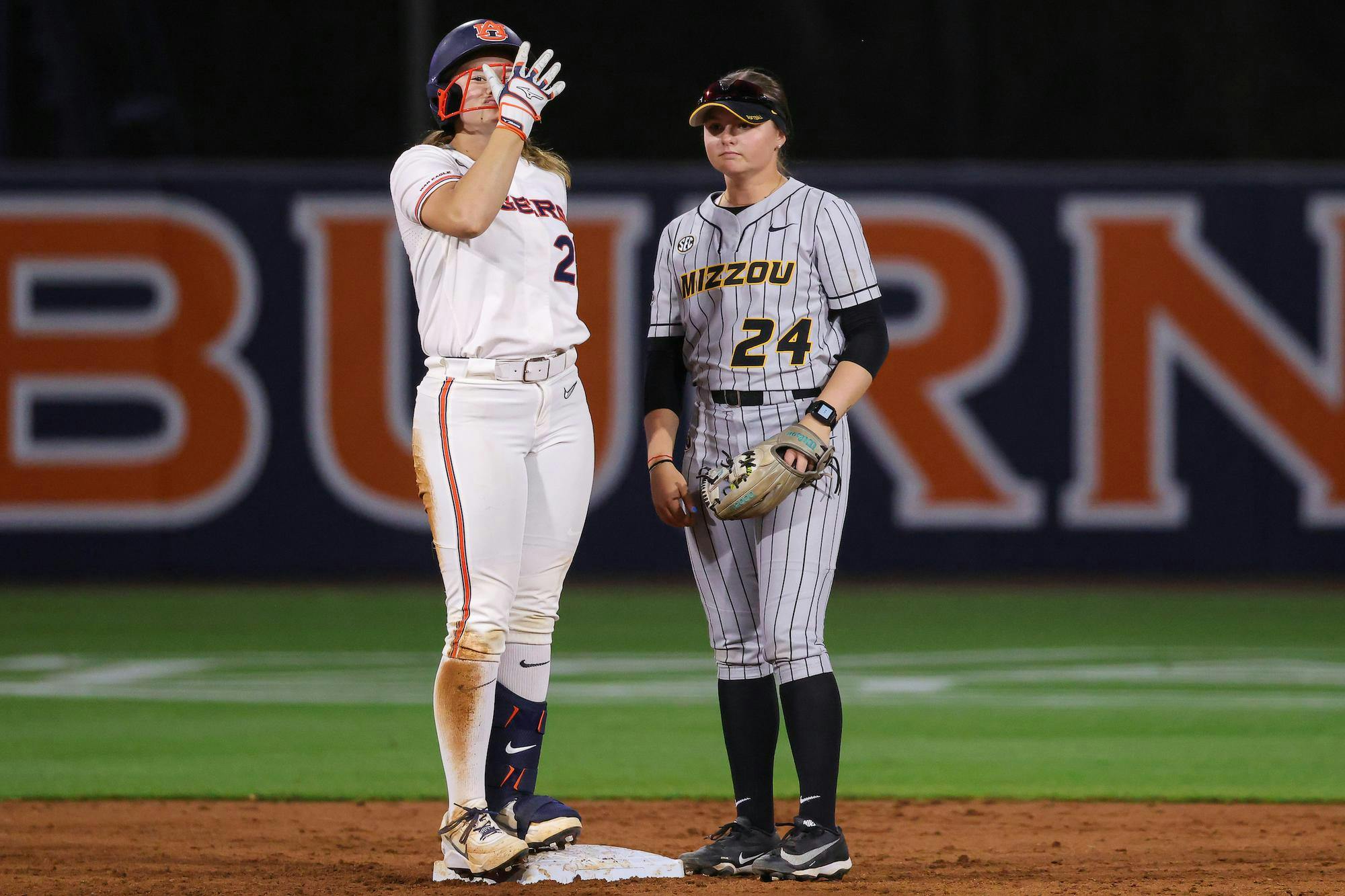 Two female softball players stand on a dirt field, one celebrating on a base while the other watches attentively.