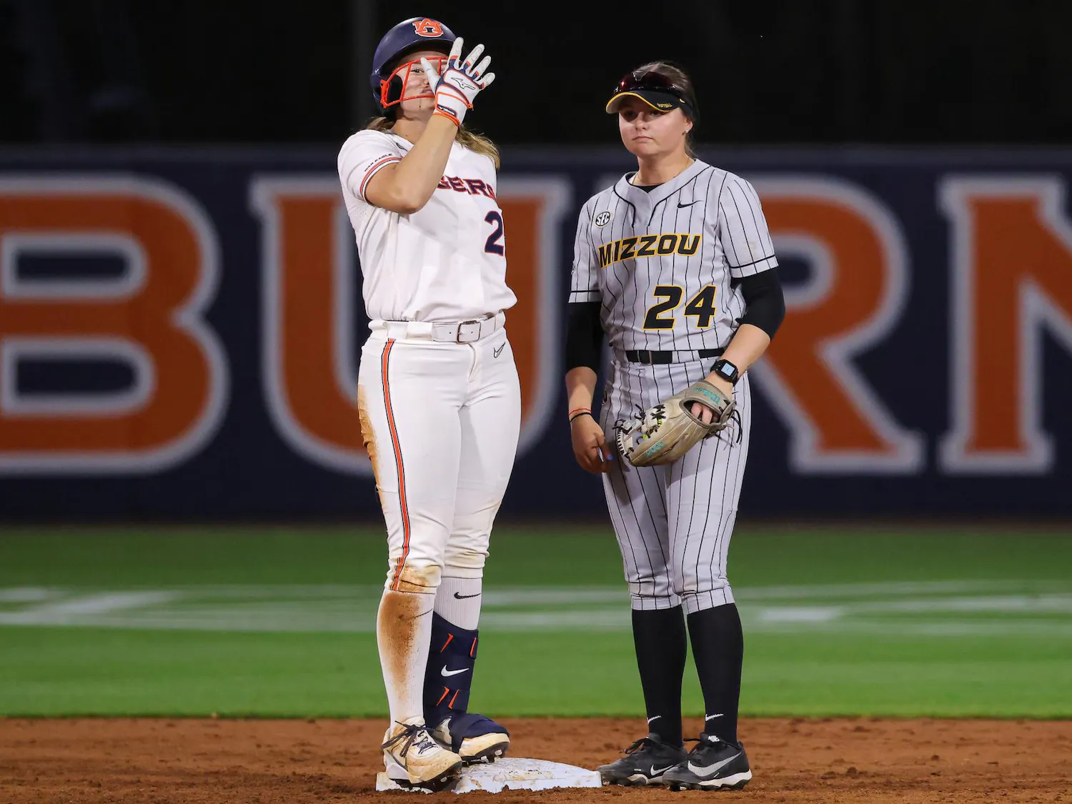 Two female softball players stand on a dirt field, one celebrating on a base while the other watches attentively.