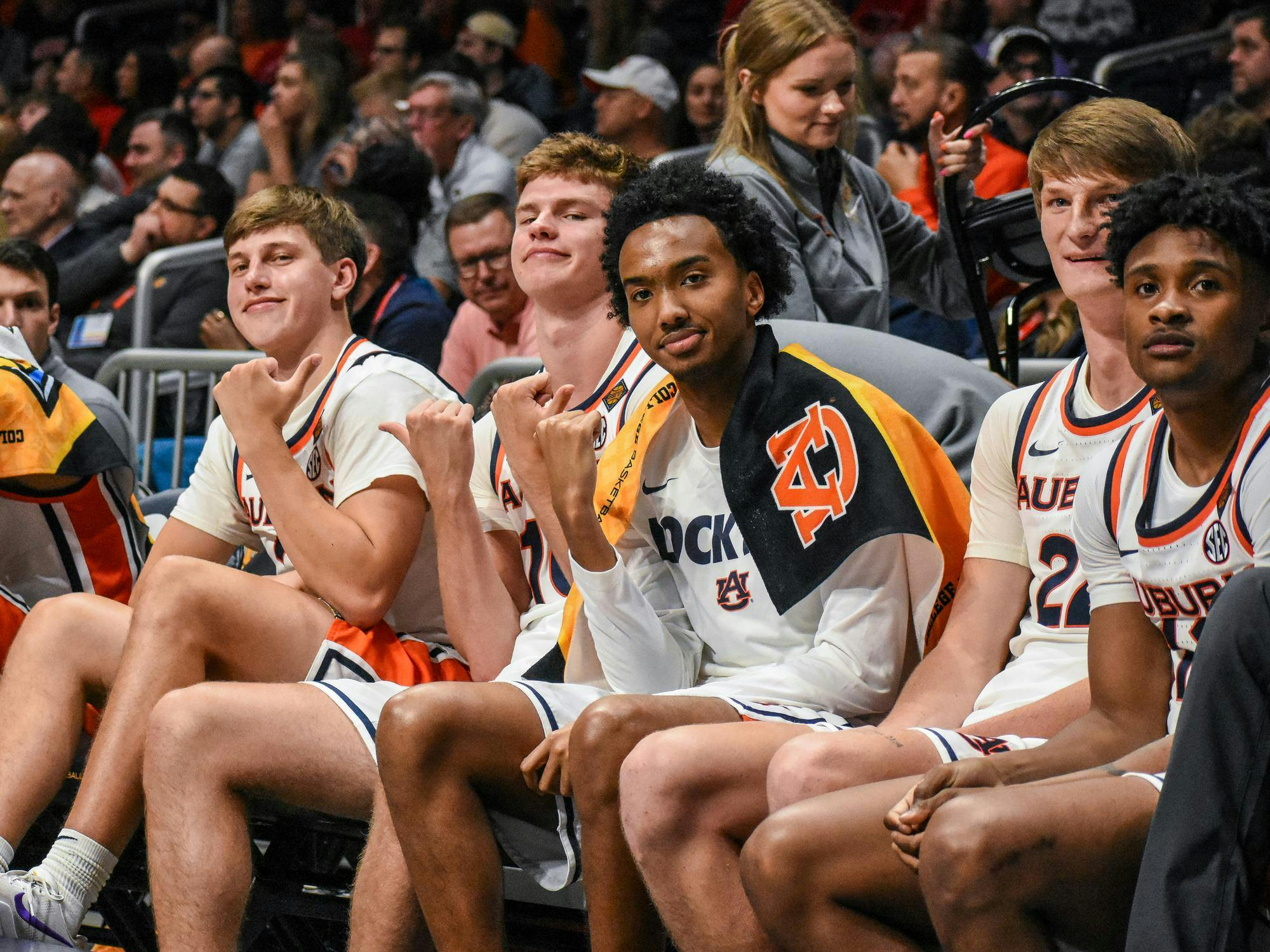 A group of six basketball players sits on a bench, smiling and making hand gestures, with fans visible in the background.