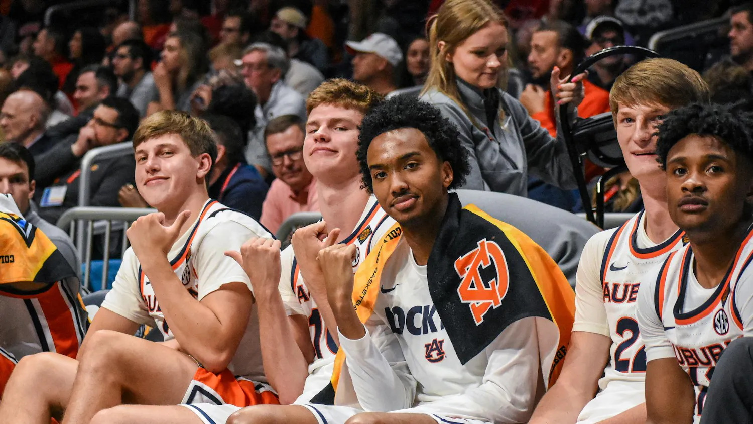 A group of six basketball players sits on a bench, smiling and making hand gestures, with fans visible in the background.