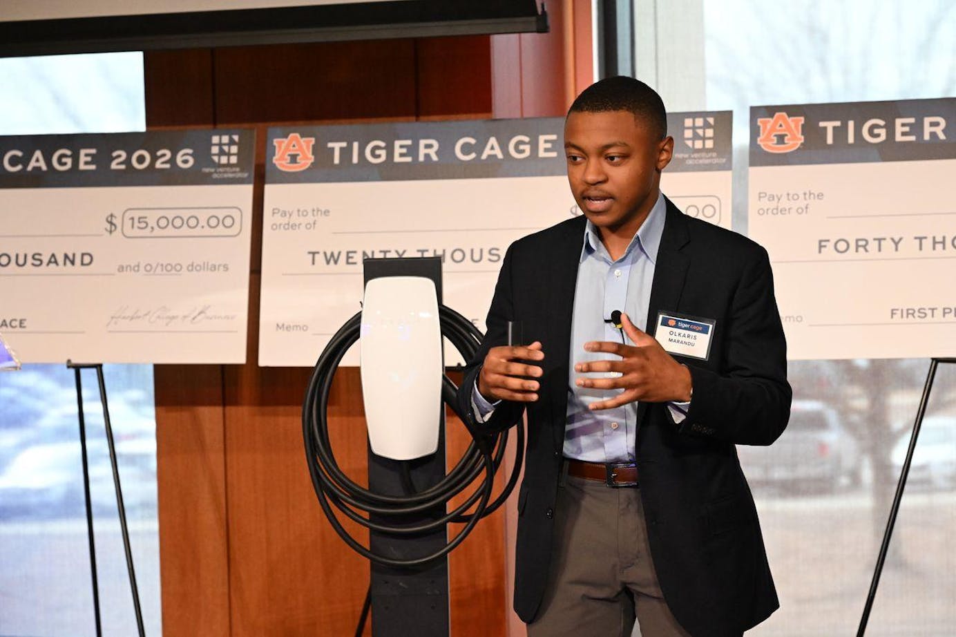 A young man in a blazer stands next to an electric vehicle charging station, gesturing while presenting in front of large checks.