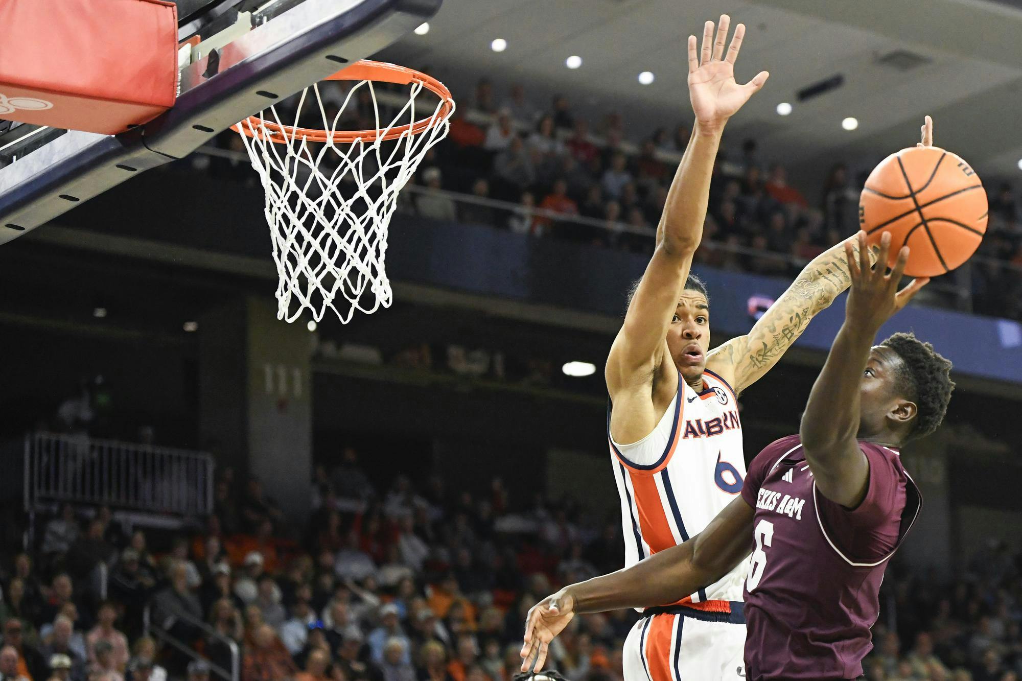 A basketball player in a maroon jersey attempts a shot while being closely guarded by an opponent in a white and orange uniform.